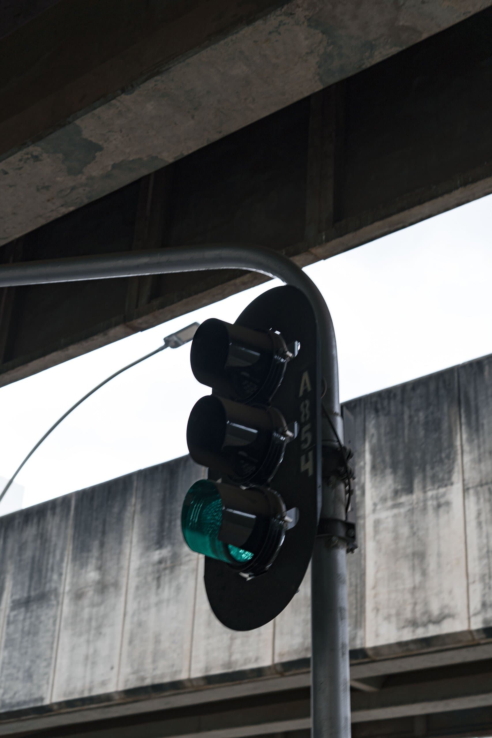A traffic light glowing green on a curved pole, against the weathered underside of an overpass.