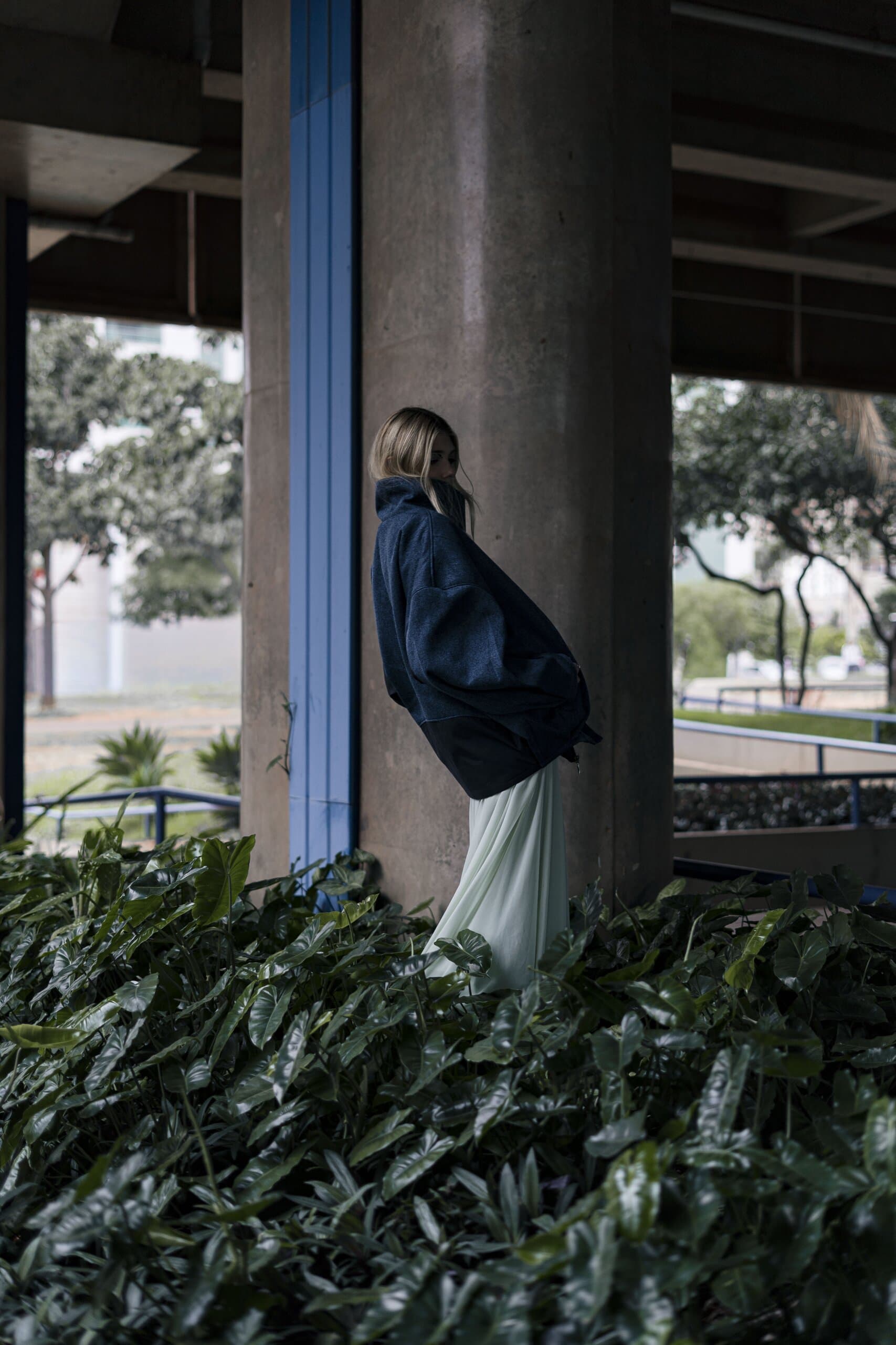 A woman in a navy coat leans against a concrete pillar striped in blue, rising out of dense green planting.