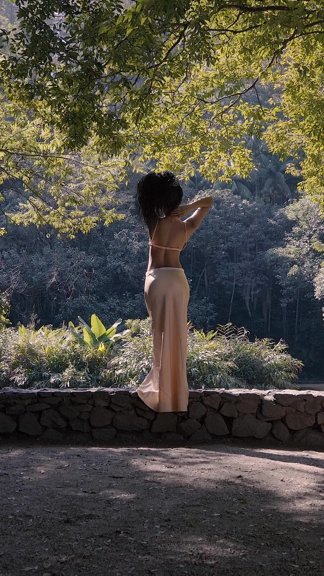 A woman in a satin skirt, seen from behind, stands on a low stone wall in dappled forest light.