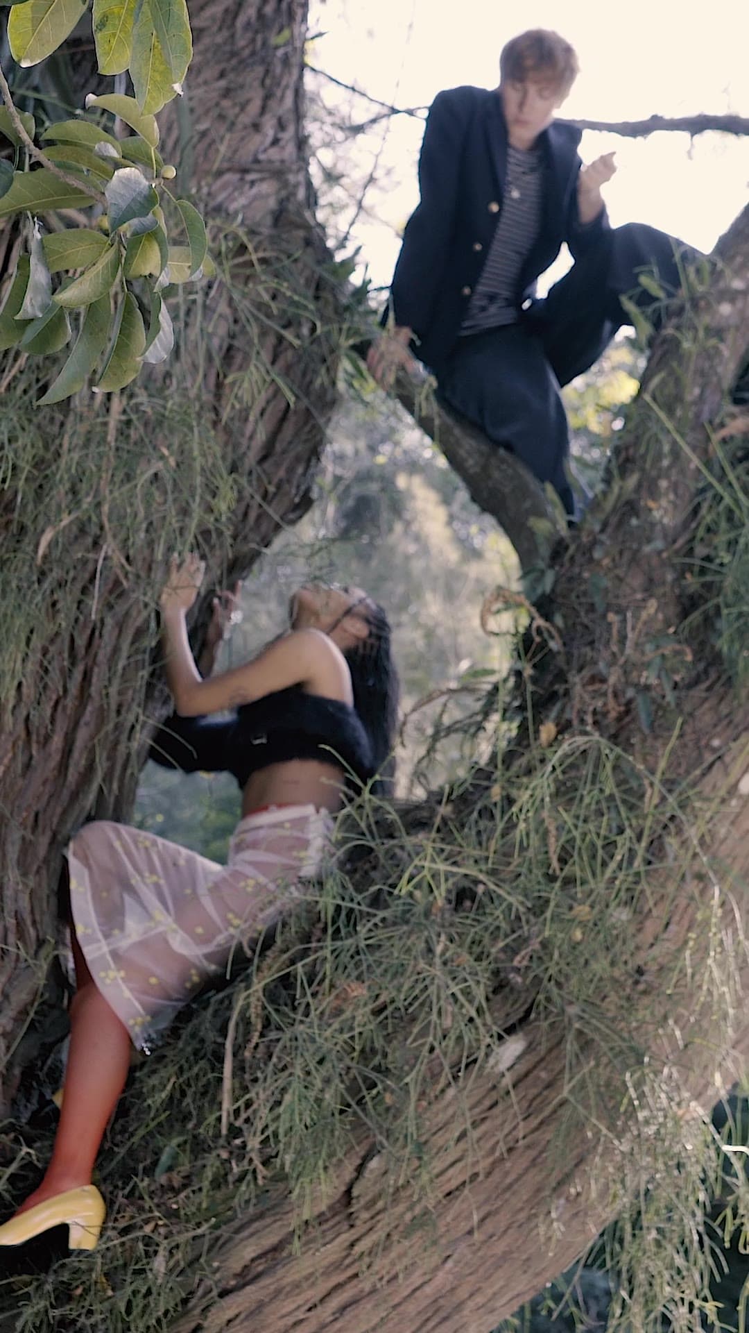 A man in a dark jacket perches in the fork of a tree while a woman leans back against the trunk below.