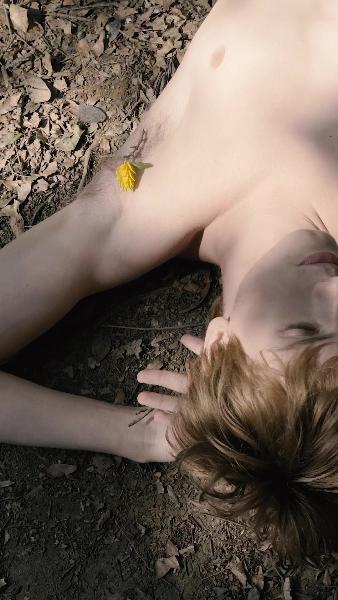 A shirtless young man lies on dry leaves and earth, a small yellow flower resting on his chest.