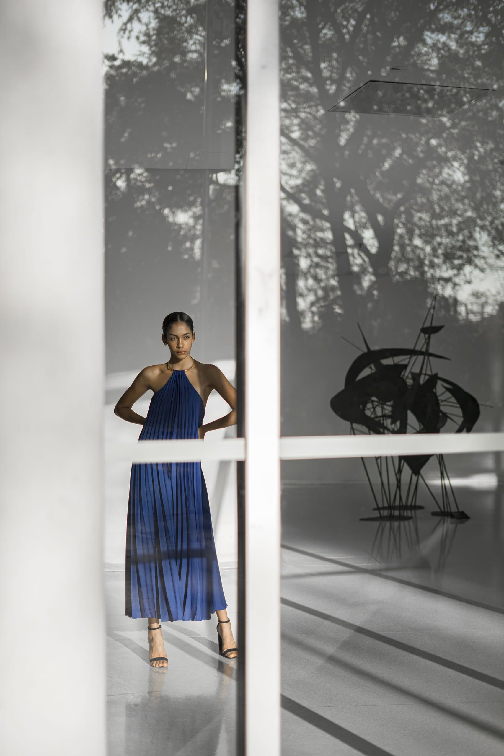 A woman in a pleated cobalt blue halter gown stands behind white window mullions, a black sculpture beside her.