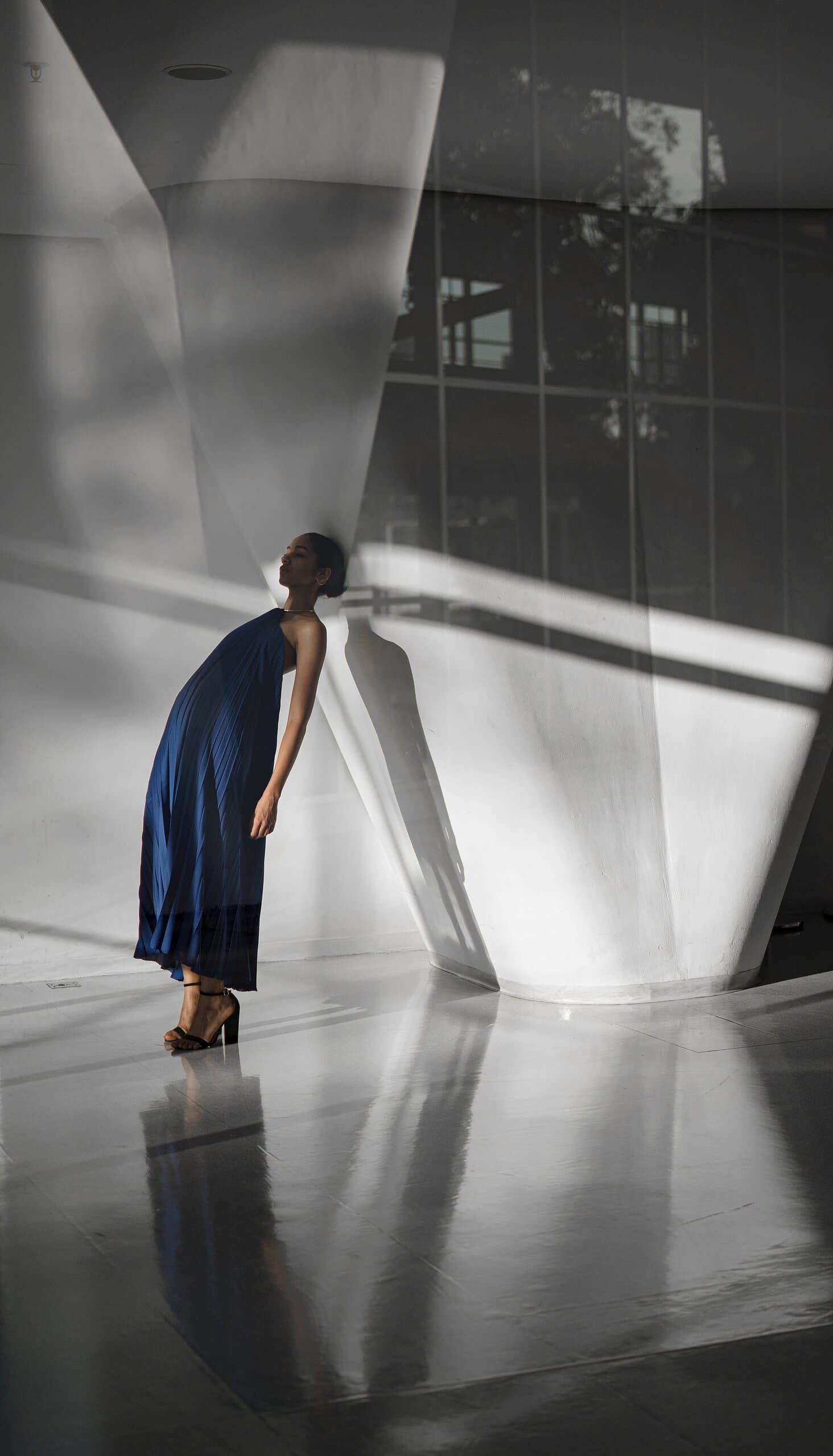 A woman in a blue gown bends forward against a white angled column, reflected in a glossy floor.