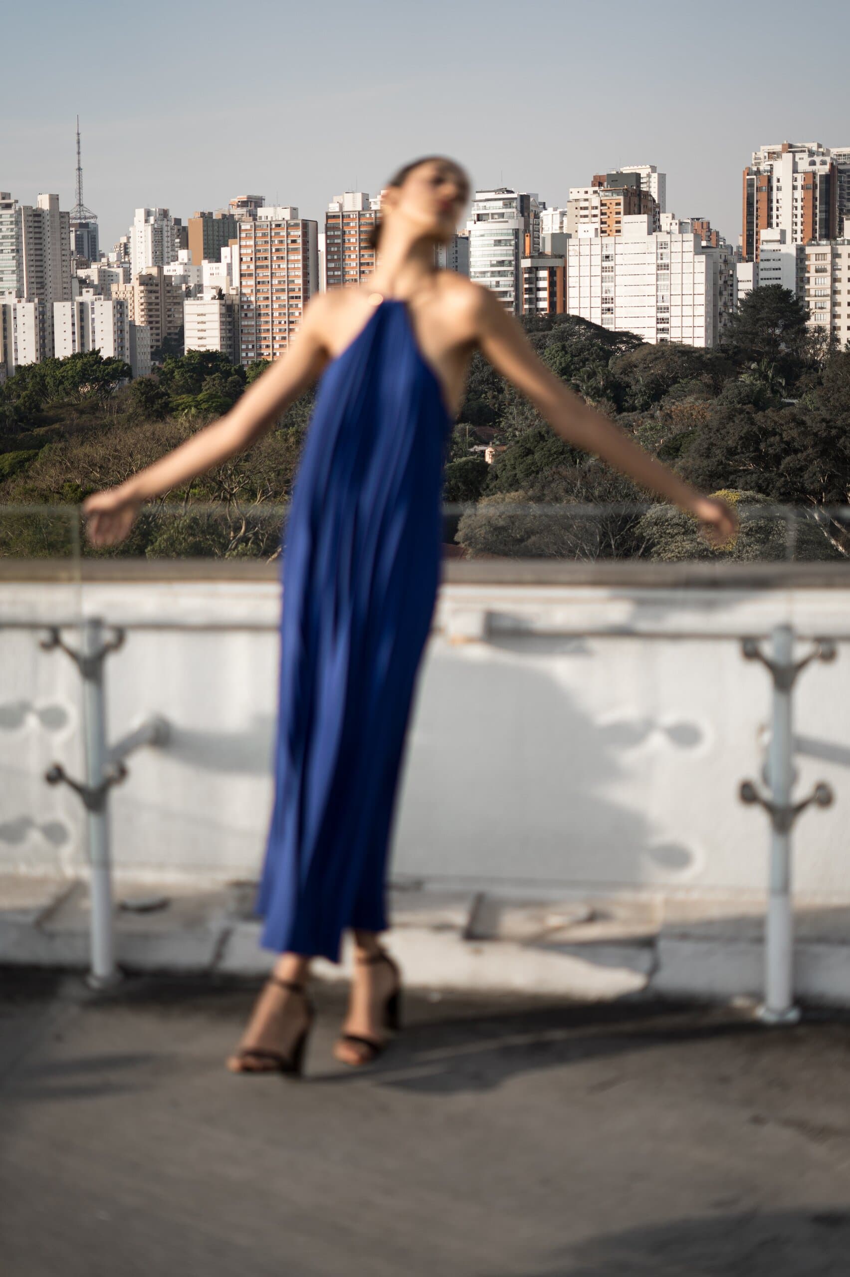 A woman in a blue halter gown flings her arms wide on a rooftop above a skyline of tower blocks.