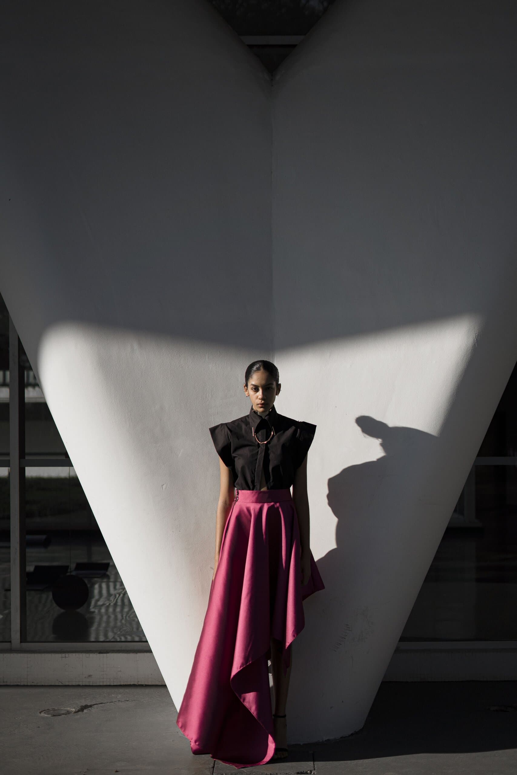 A woman in a black sleeveless top and a magenta draped skirt stands before a white V-shaped concrete column.