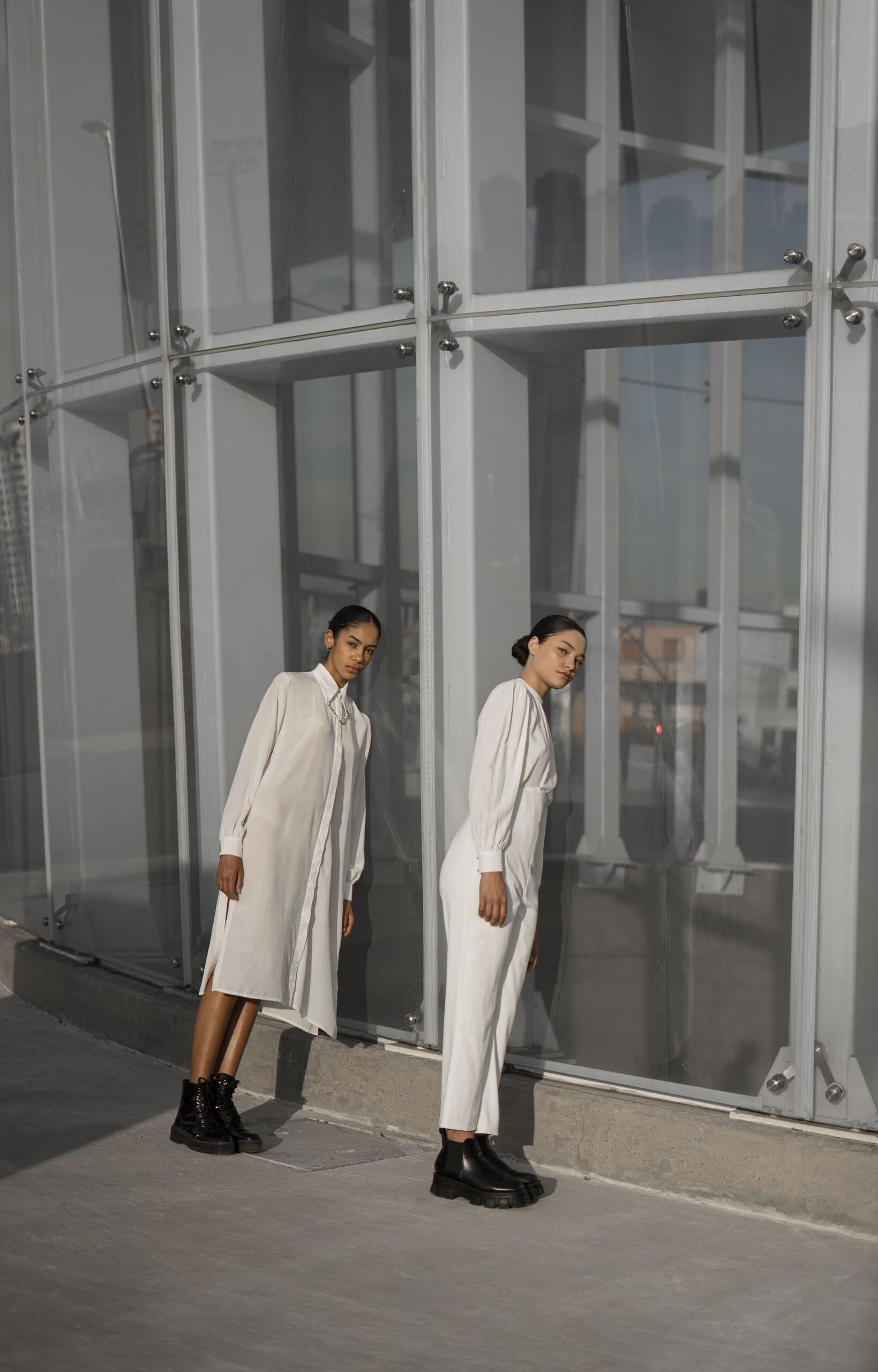 Two women in white lean against a curved glass wall, one in a long shirt dress and one in a jumpsuit.