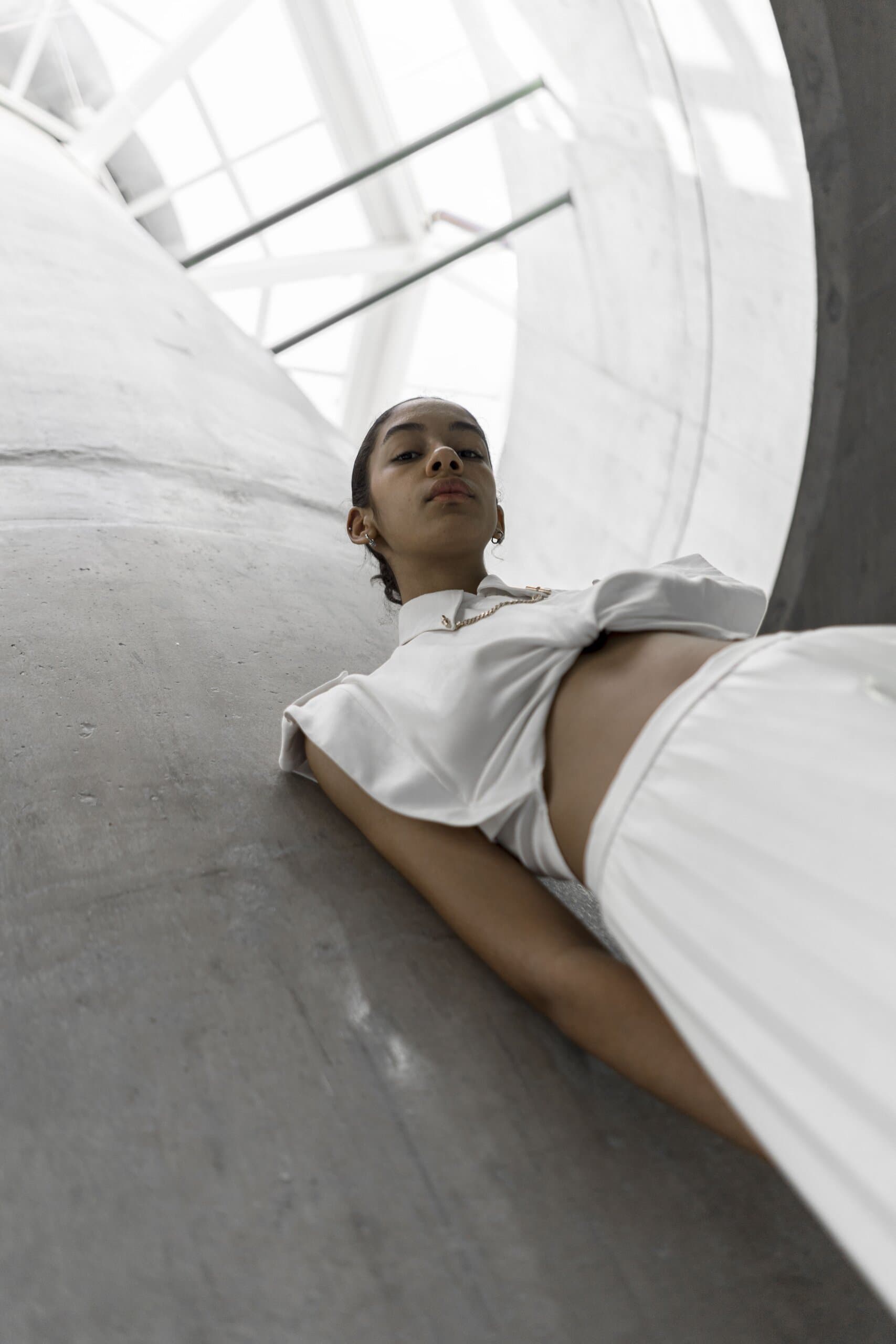 From below: a woman in a white cropped top lies back against curved concrete under a glazed roof.