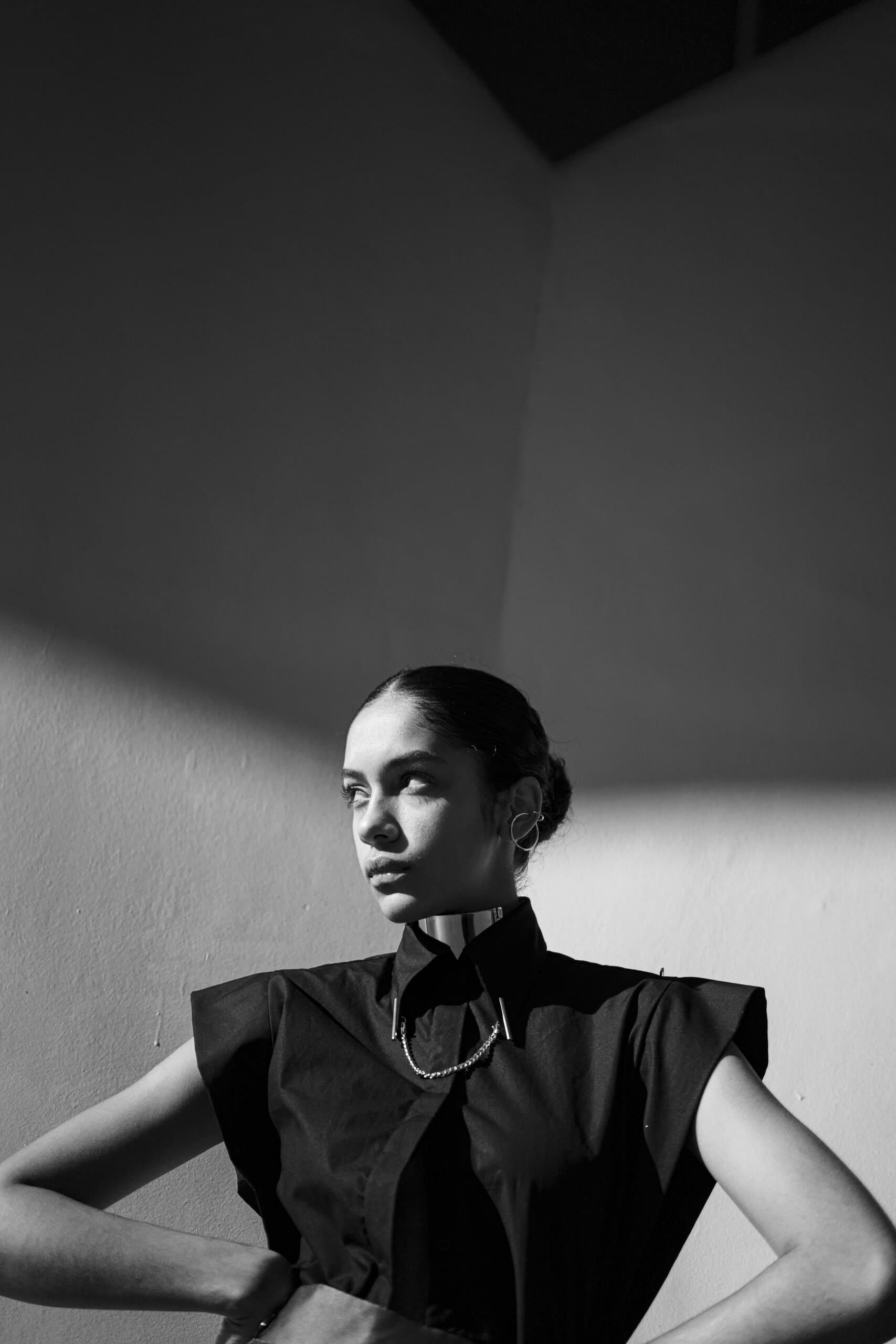 Black and white: a woman in a black sleeveless shirt with a high collar, hands on her hips, half in shadow.