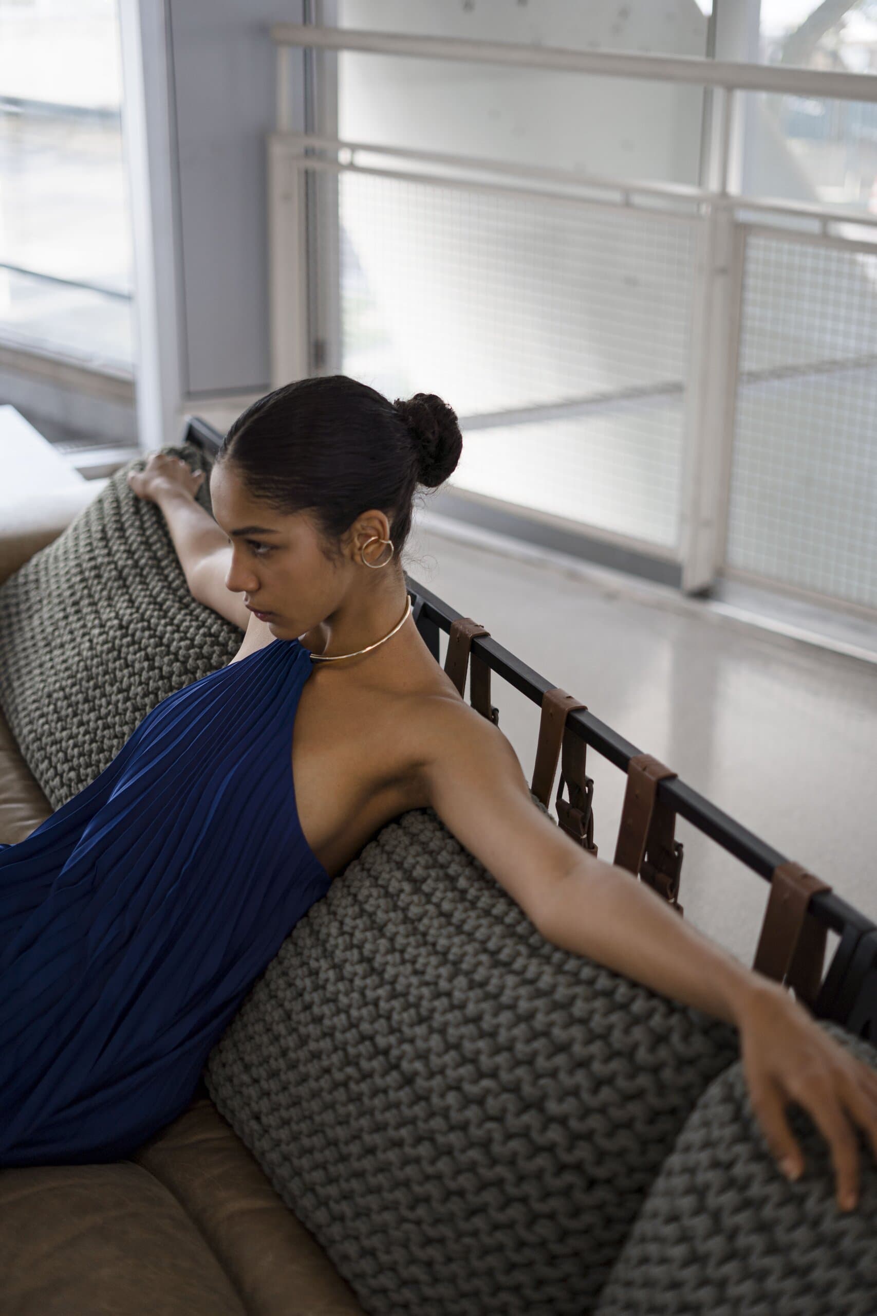 A woman in a cobalt blue one-shoulder dress leans across a woven grey sofa by a bright window.