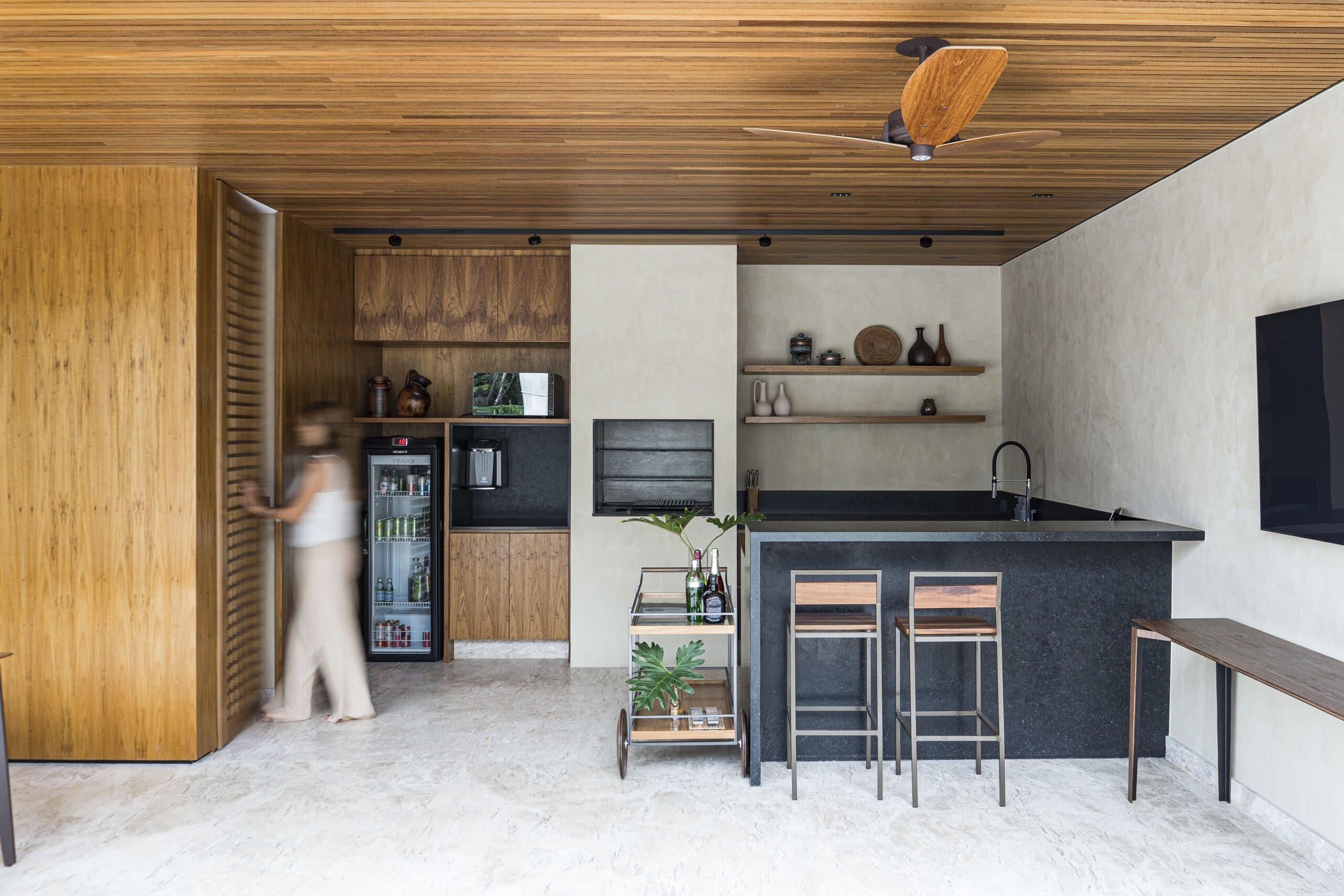 An outdoor kitchen under a timber ceiling: a dark bar with two stools, open shelves and a figure blurred at the door.