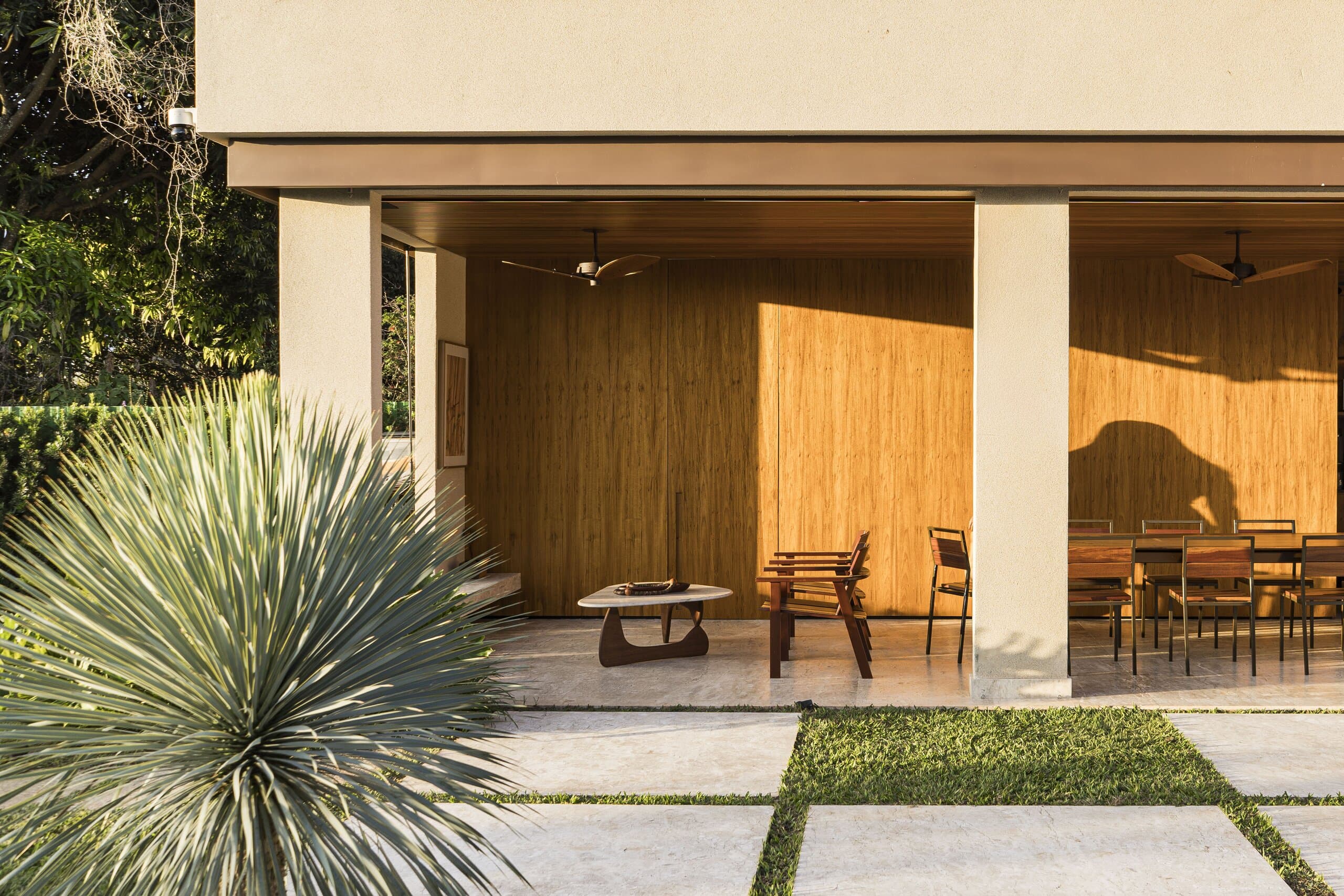 A deep veranda in sunlight: timber walls, dining chairs and a spiky agave in the garden in front.