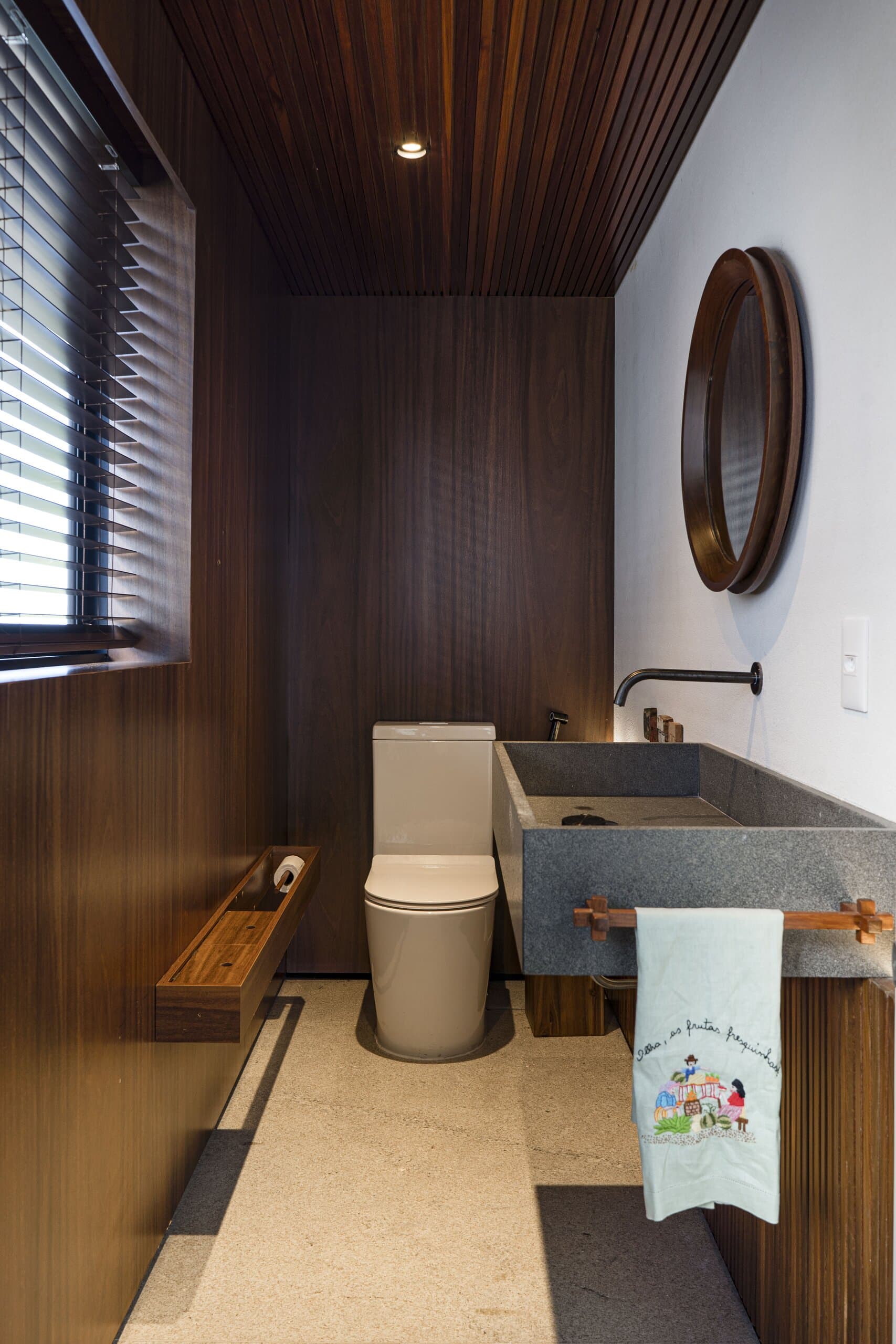Um lavabo revestido de madeira escura, com cuba de pedra, espelho redondo e uma toalha bordada.