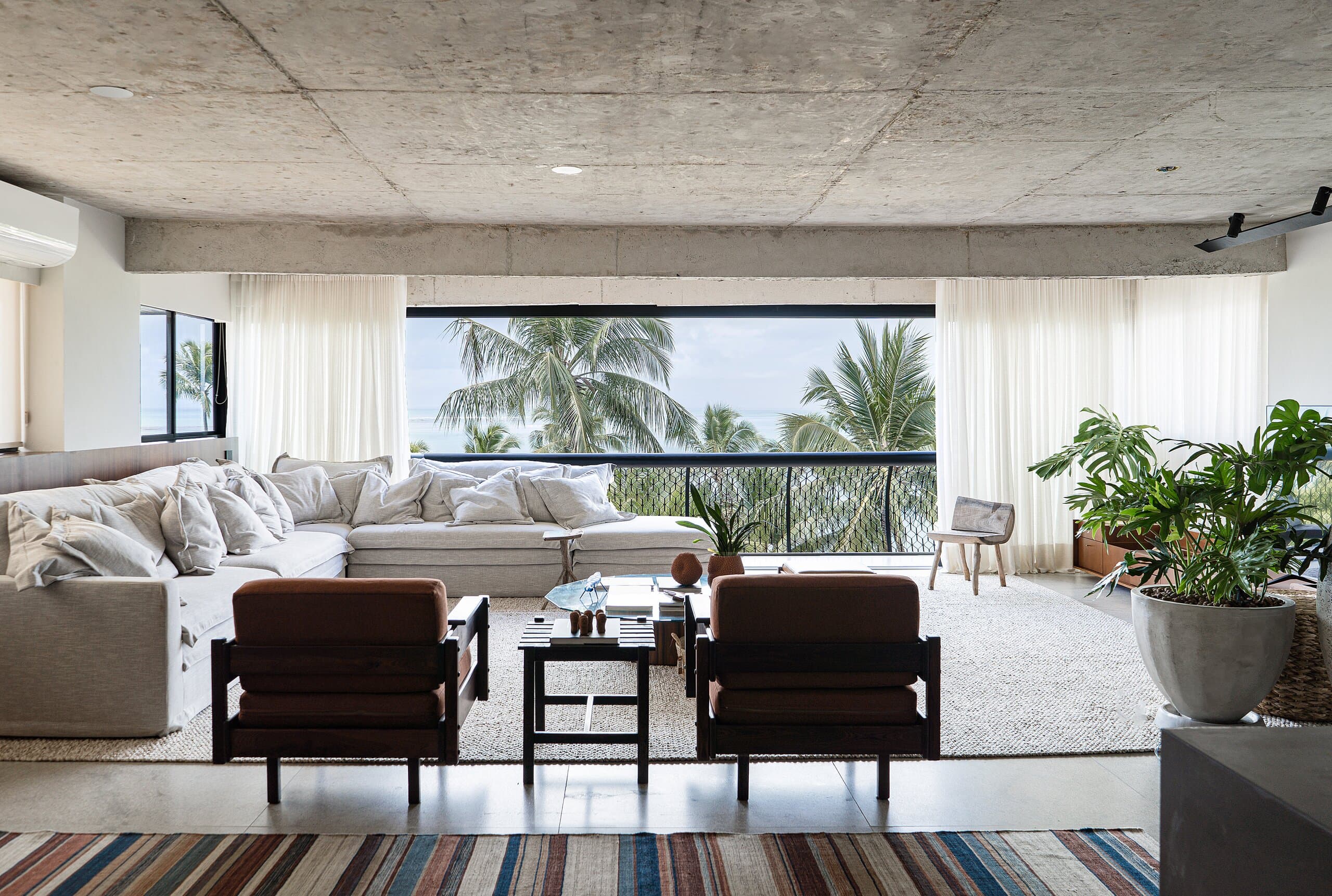 A sala vista por cima de um tapete listrado: duas poltronas de couro, um longo sofá branco e coqueiros além do vidro da varanda.