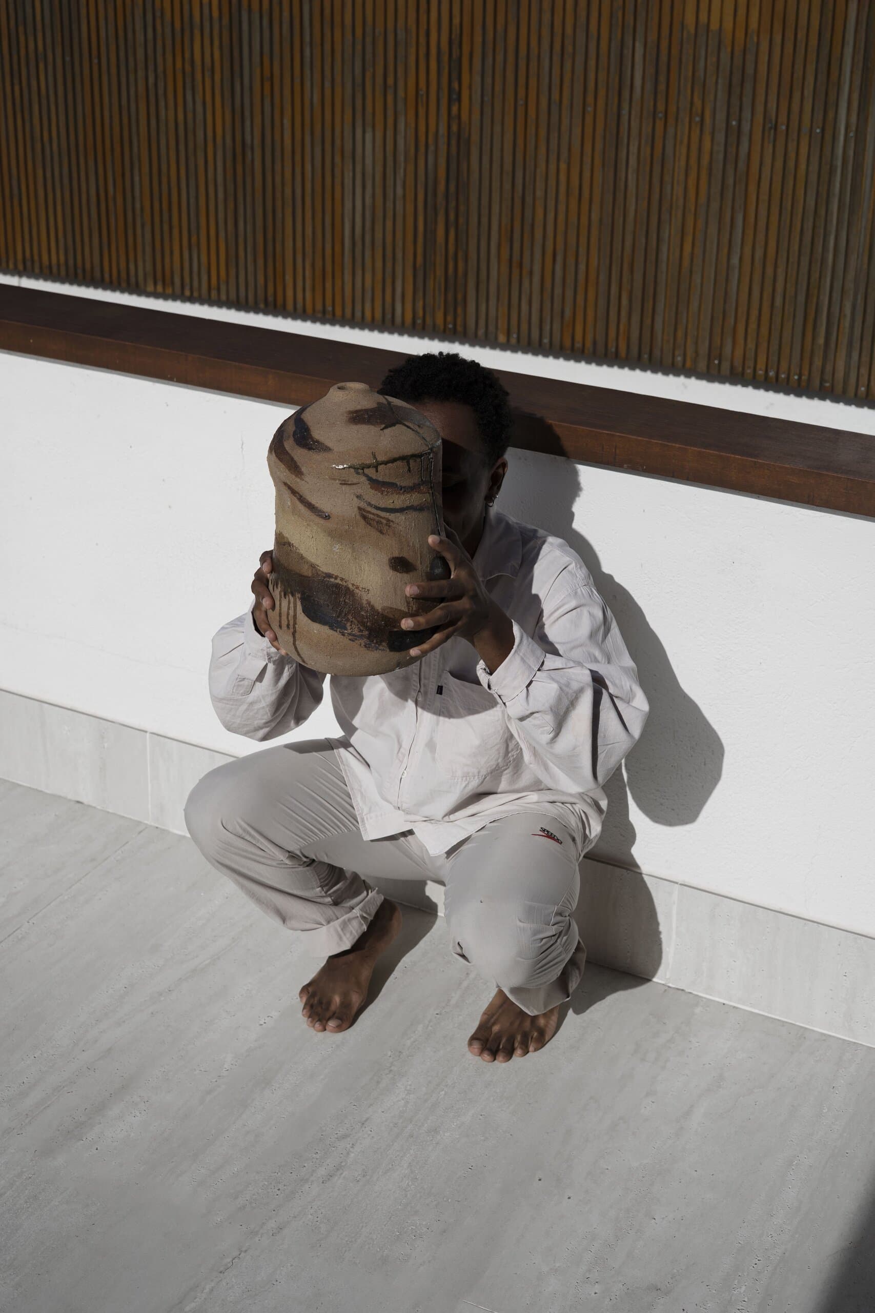 A man in white crouches barefoot against a white wall, holding a large clay vessel over his face.