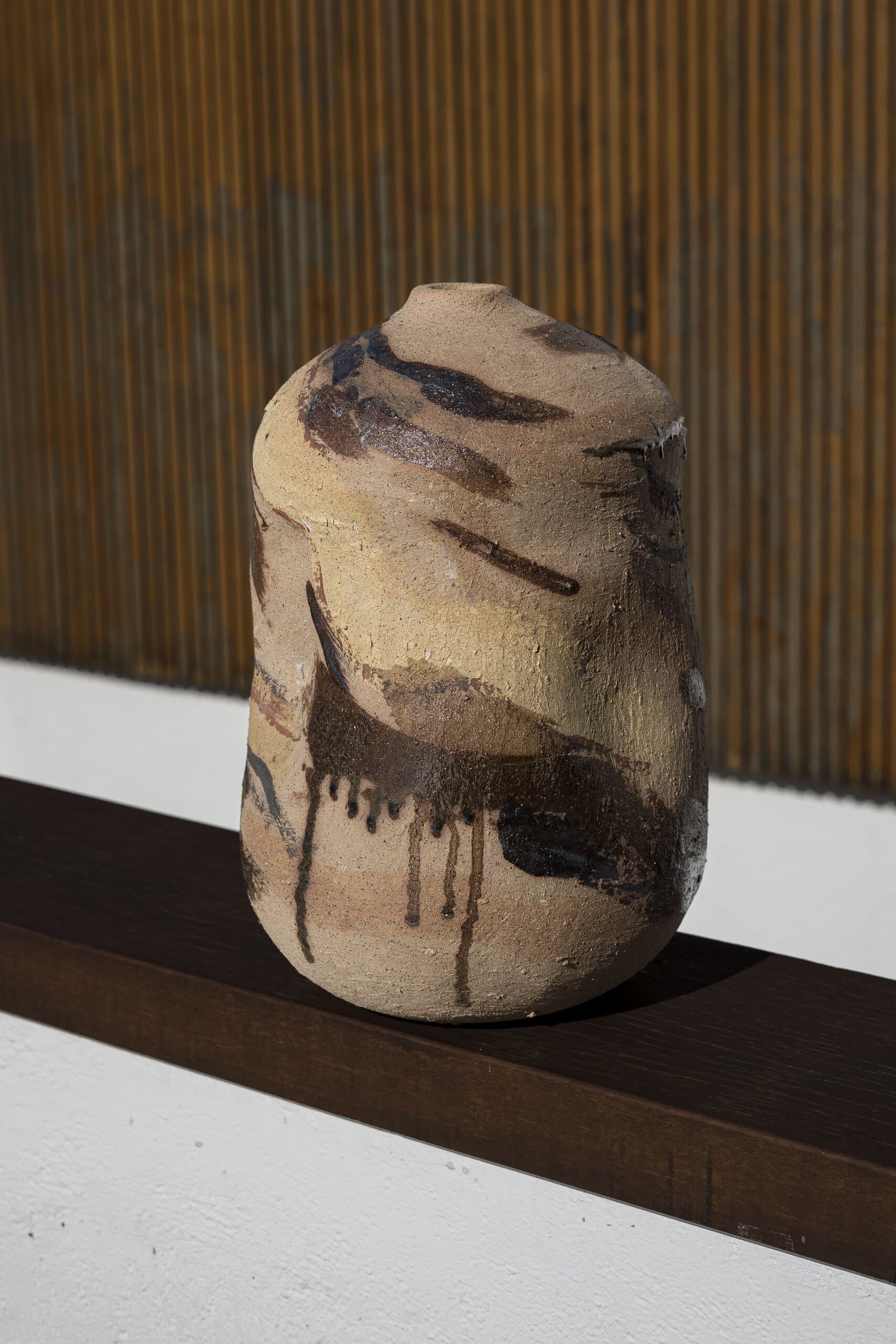 A tall vessel streaked with dark glaze drips on a timber ledge, slatted wood behind.