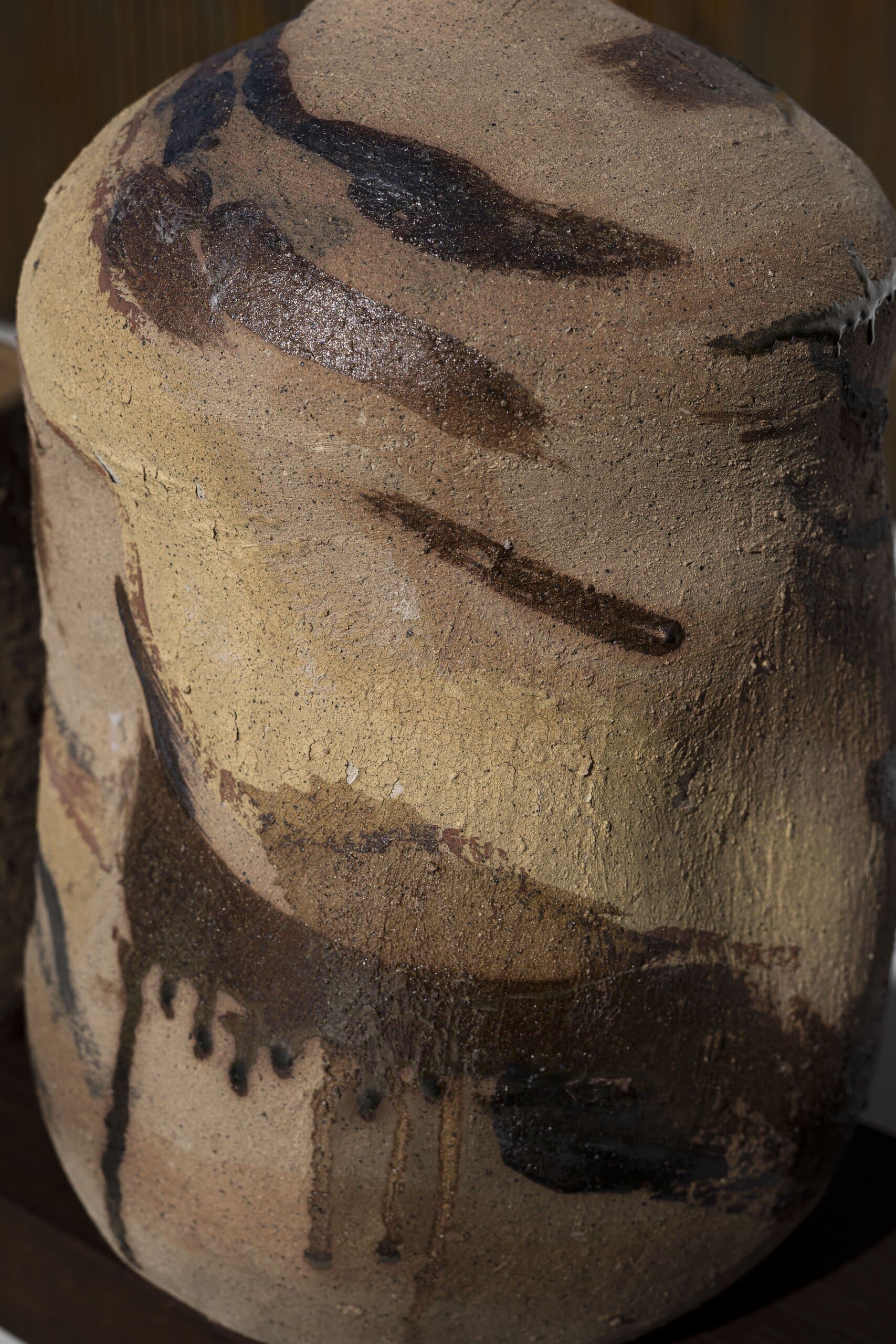 Close up: the sandy surface of a vessel, brushed with dark glaze that runs down in drips.