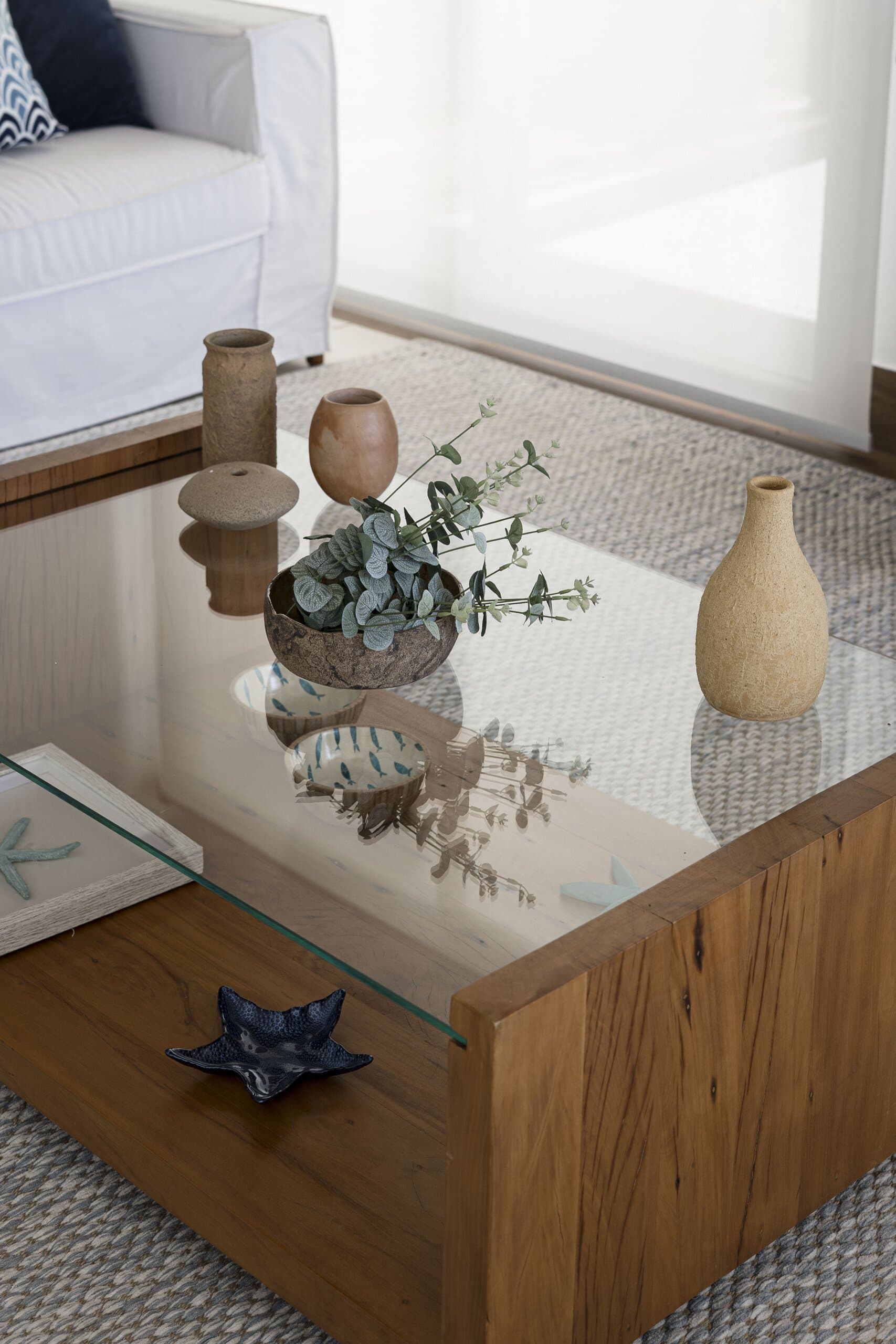 Clay vases and a sprig of eucalyptus on a glass coffee table with a solid timber base, beside a white sofa.