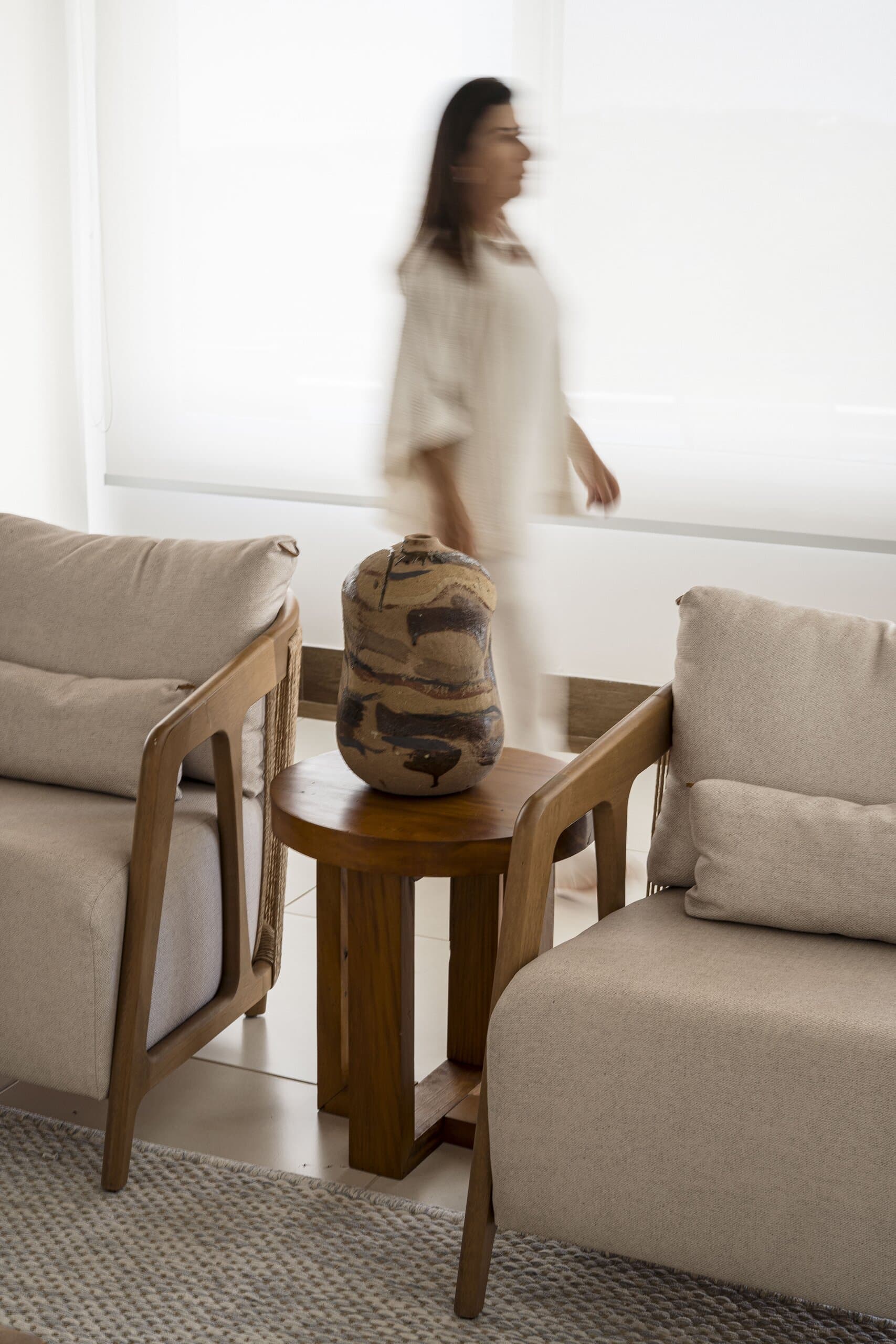 A glazed vessel on a timber side table between two linen armchairs, a woman in white blurred as she passes.