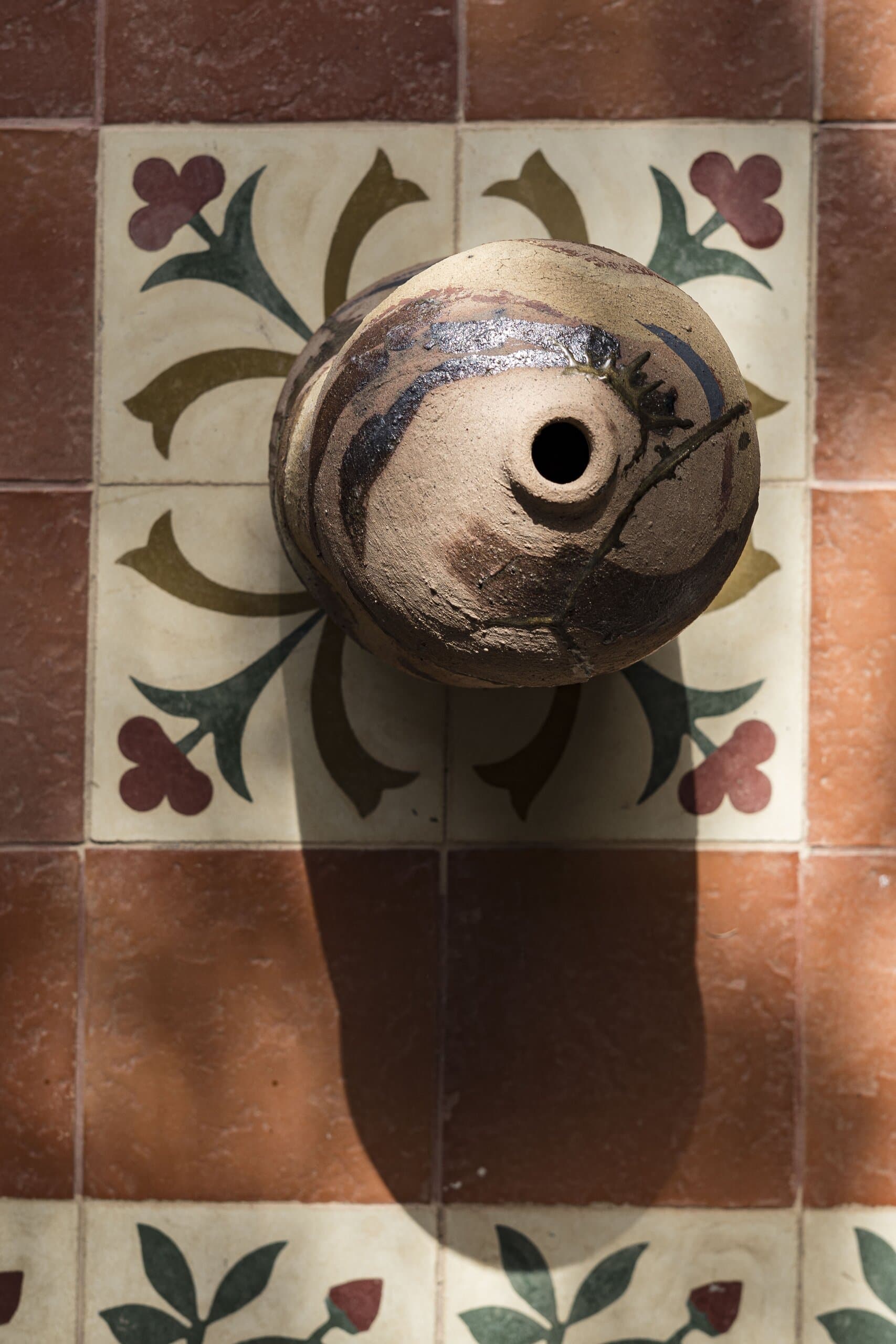 From above: the open mouth of a glazed vessel on a tile of painted leaves, its shadow falling across terracotta.