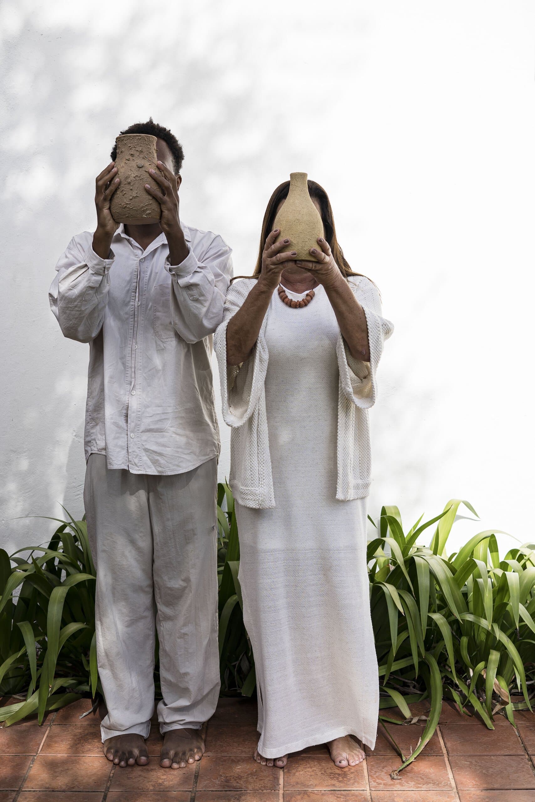 A man and a woman in white linen hold ceramic vessels over their faces against a white wall and green leaves.