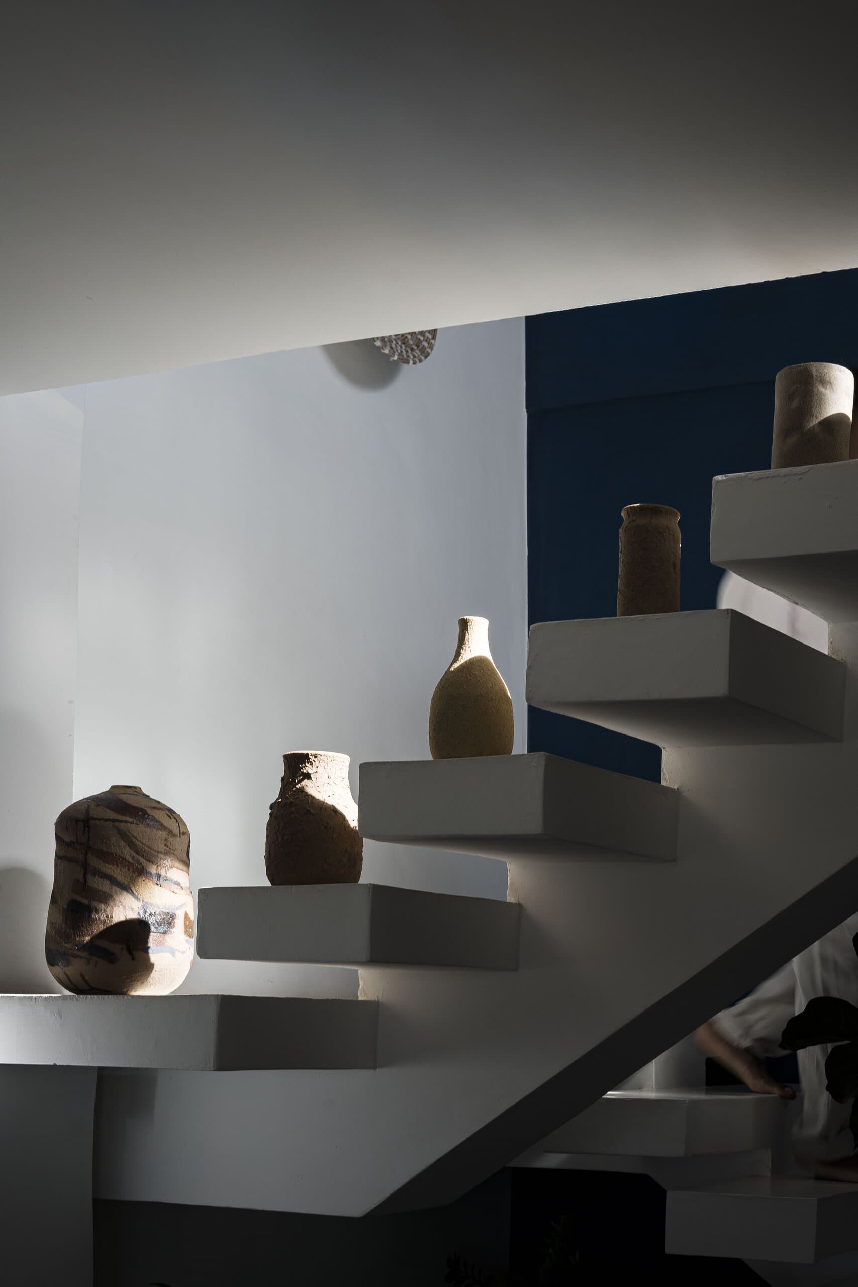 Clay vessels set on the treads of a white floating staircase, in raking light.