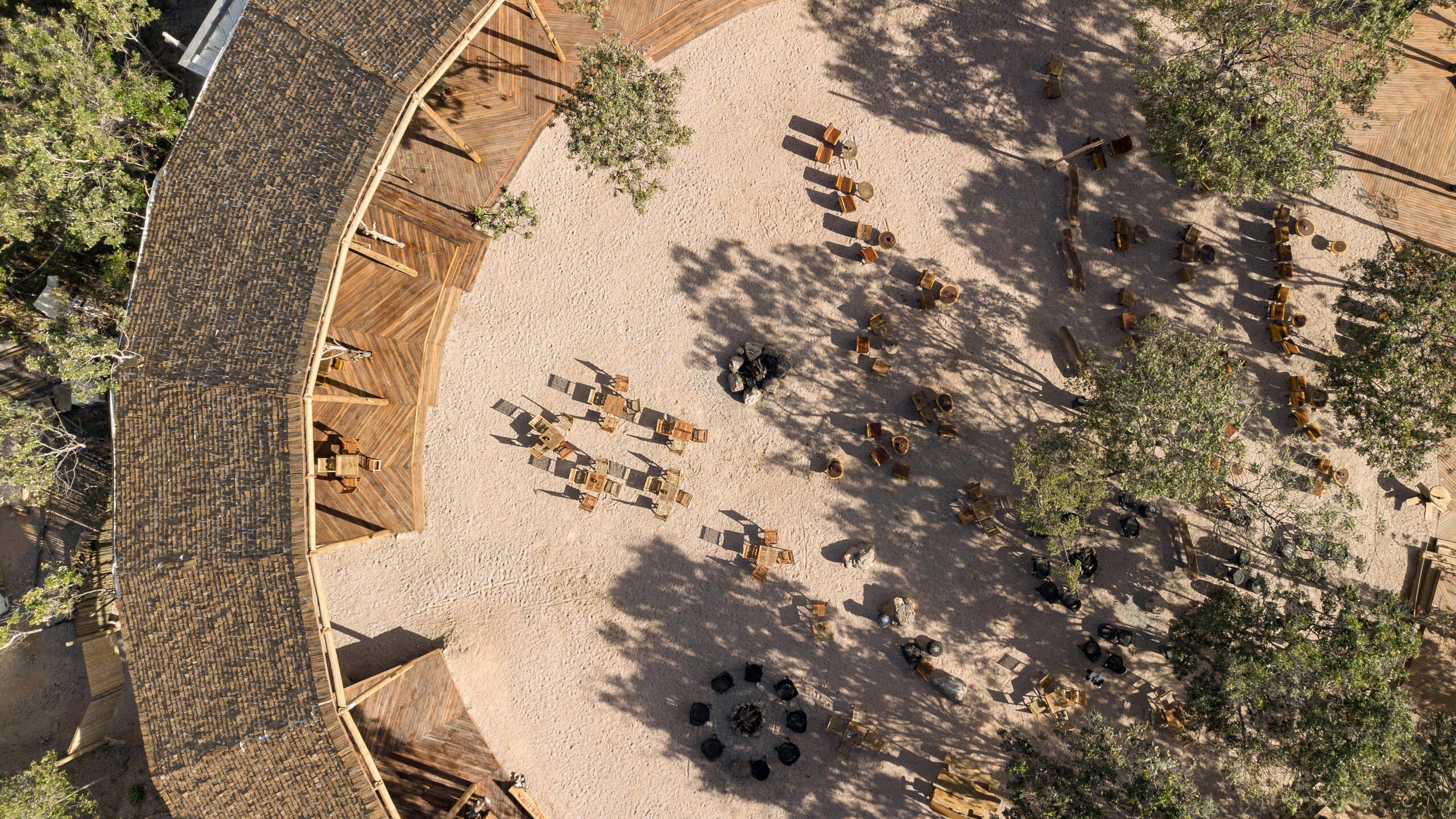 Overhead: the curve of the thatched roof beside a sandy courtyard of tables, chairs and trees casting shadows.