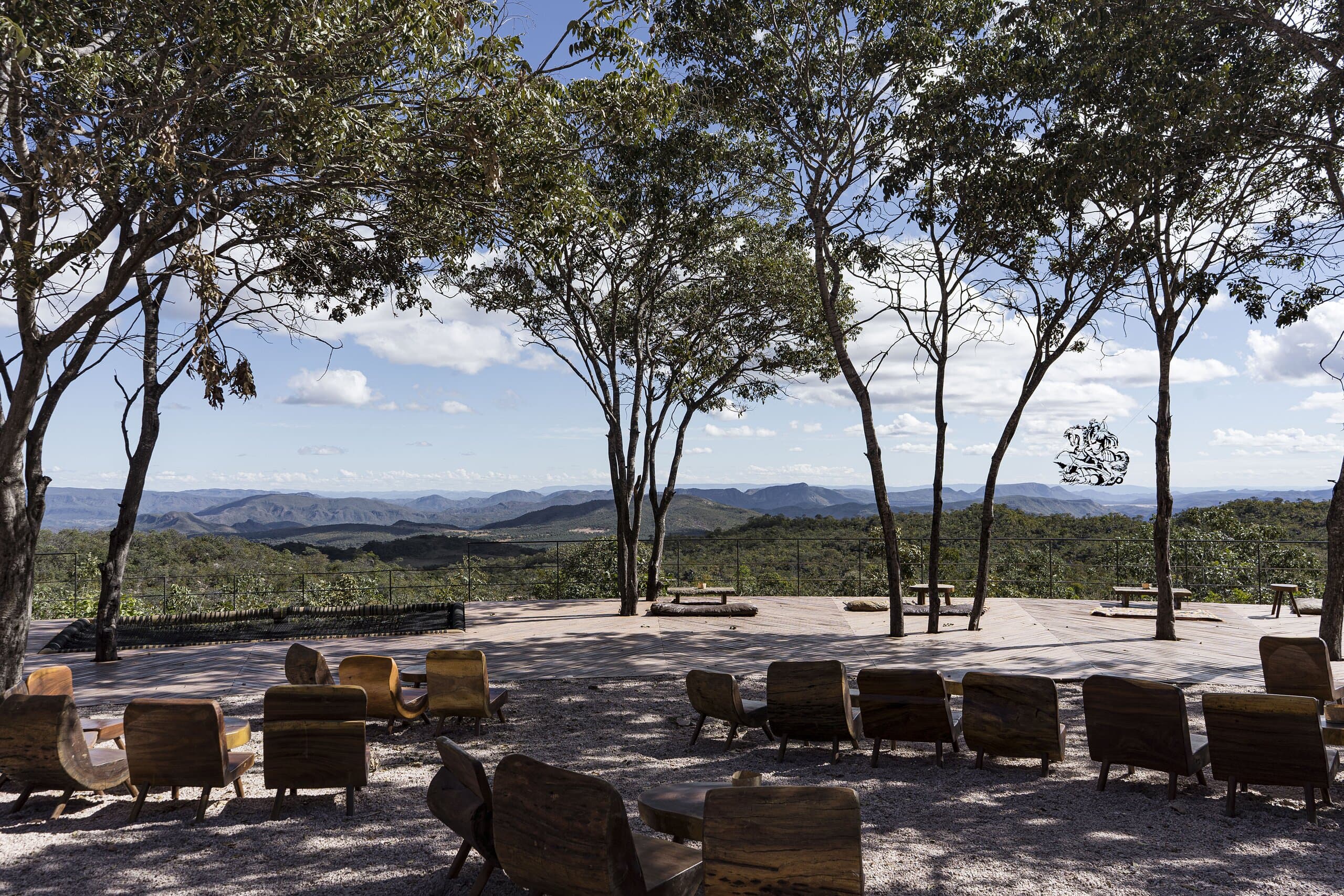 Armchairs scattered on a sandy terrace under slender trees, hills fading into the blue distance.