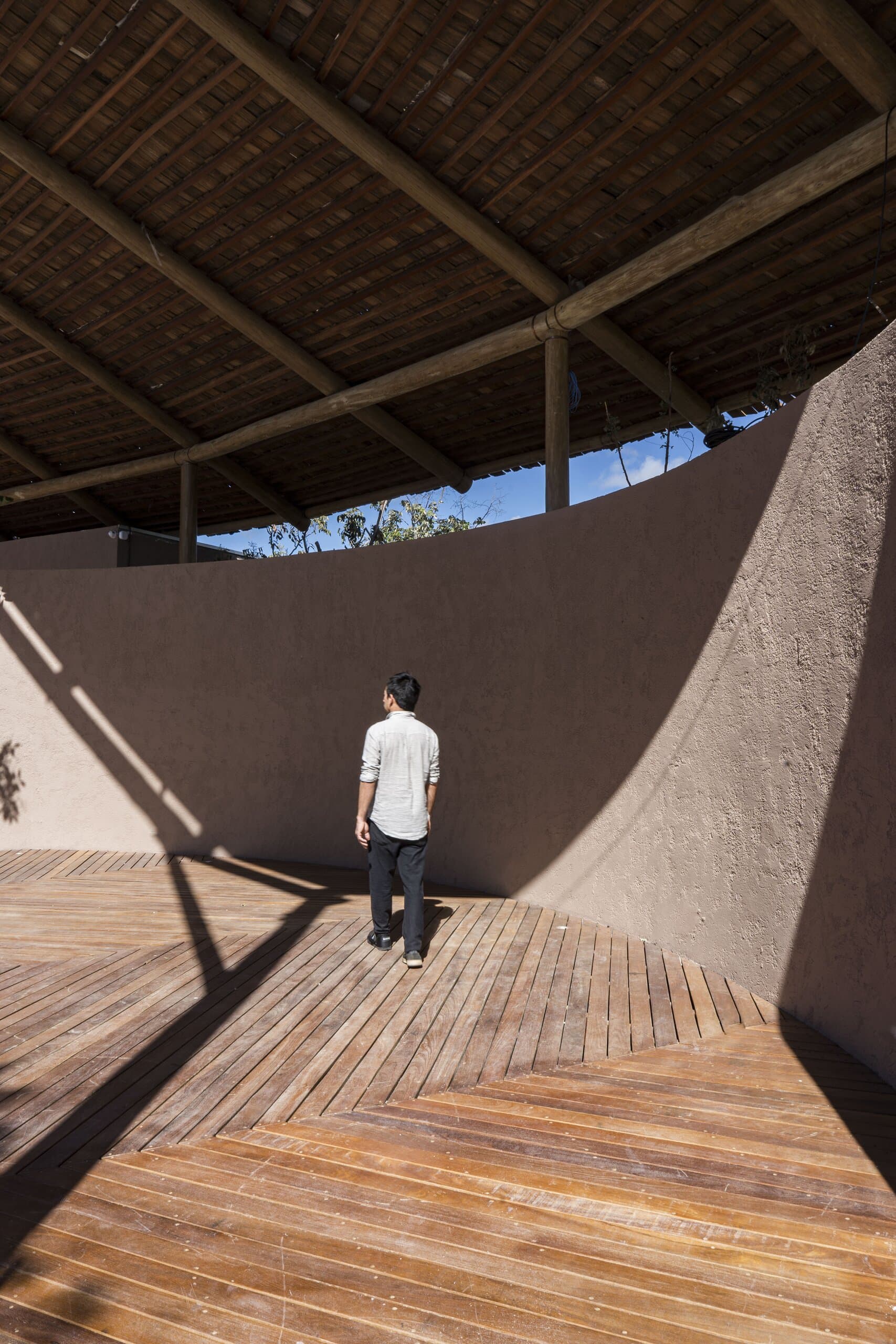 A man in white stands on a timber deck before a curved earthen wall beneath the slatted roof.