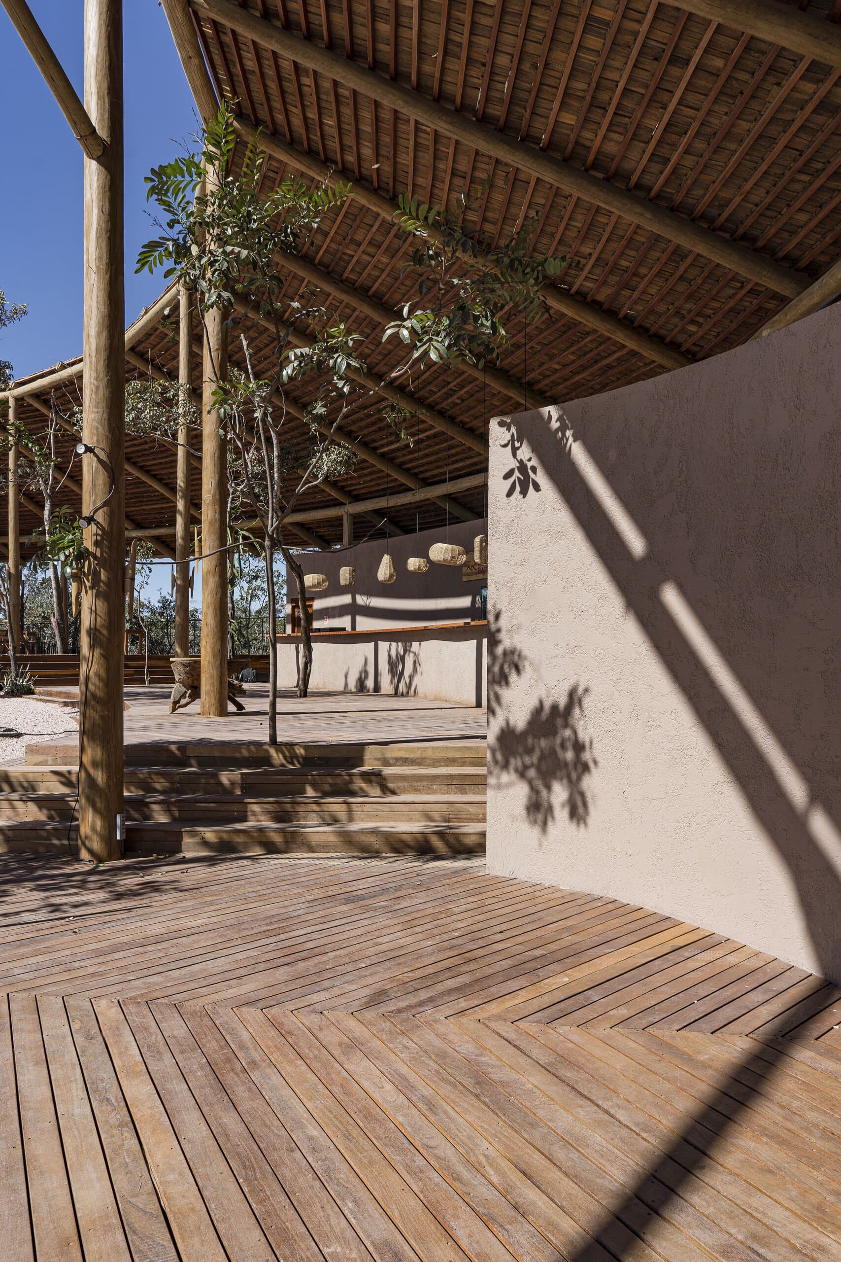 Shadows of a tree on a curved earthen wall, timber posts and a slatted roof above the deck.