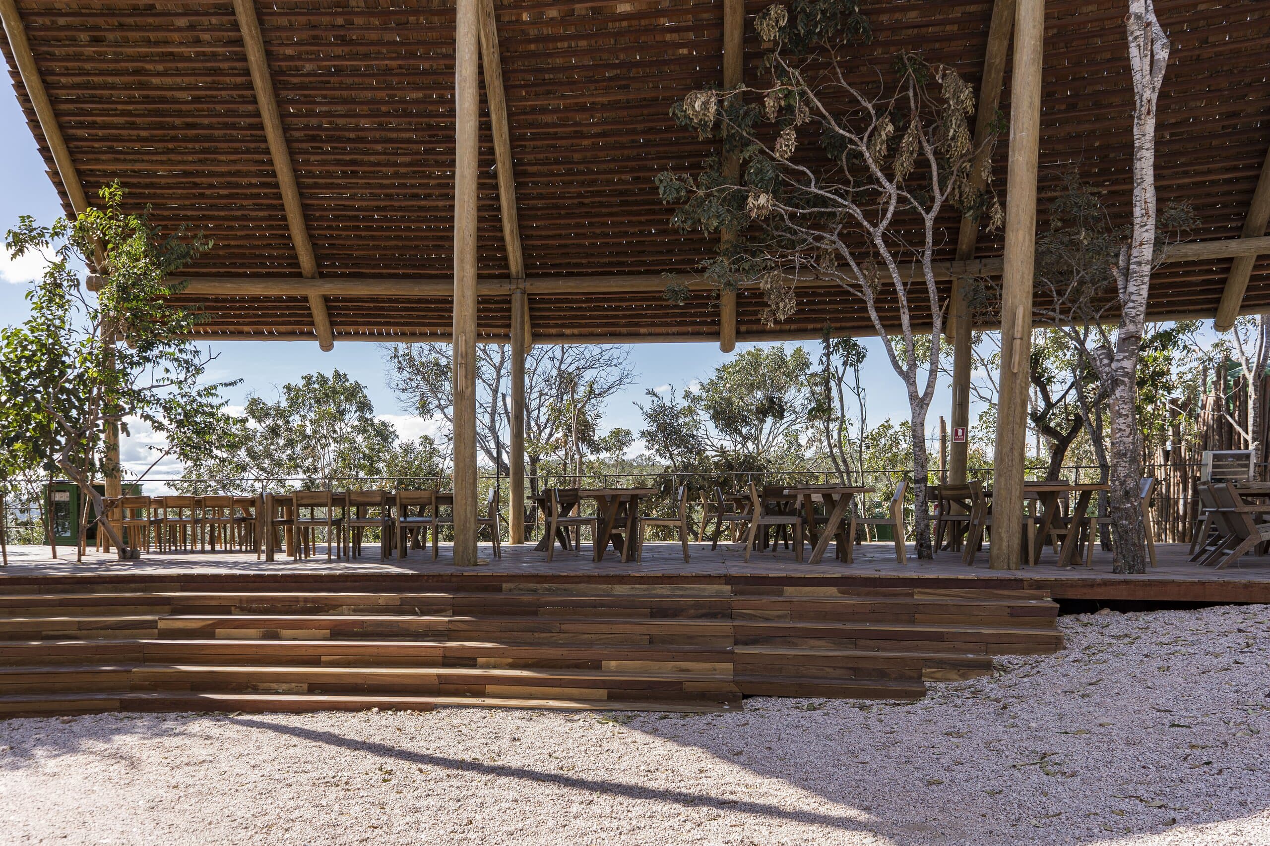 Timber steps rise from the sand to a deck under the wide slatted roof, trees growing through it.