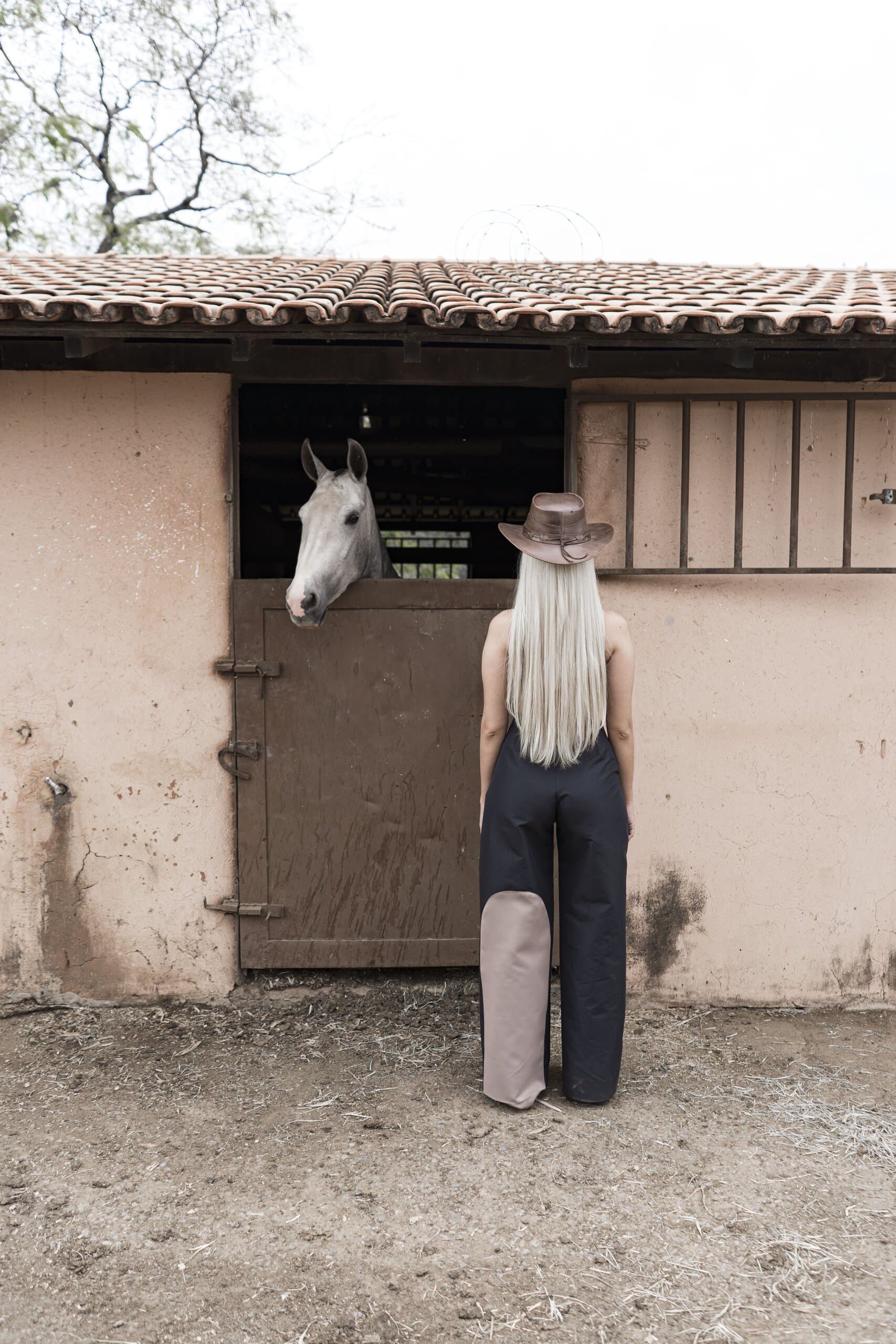 A platinum-haired woman in a leather hat faces a pink stable wall as a white horse looks out of its stall.