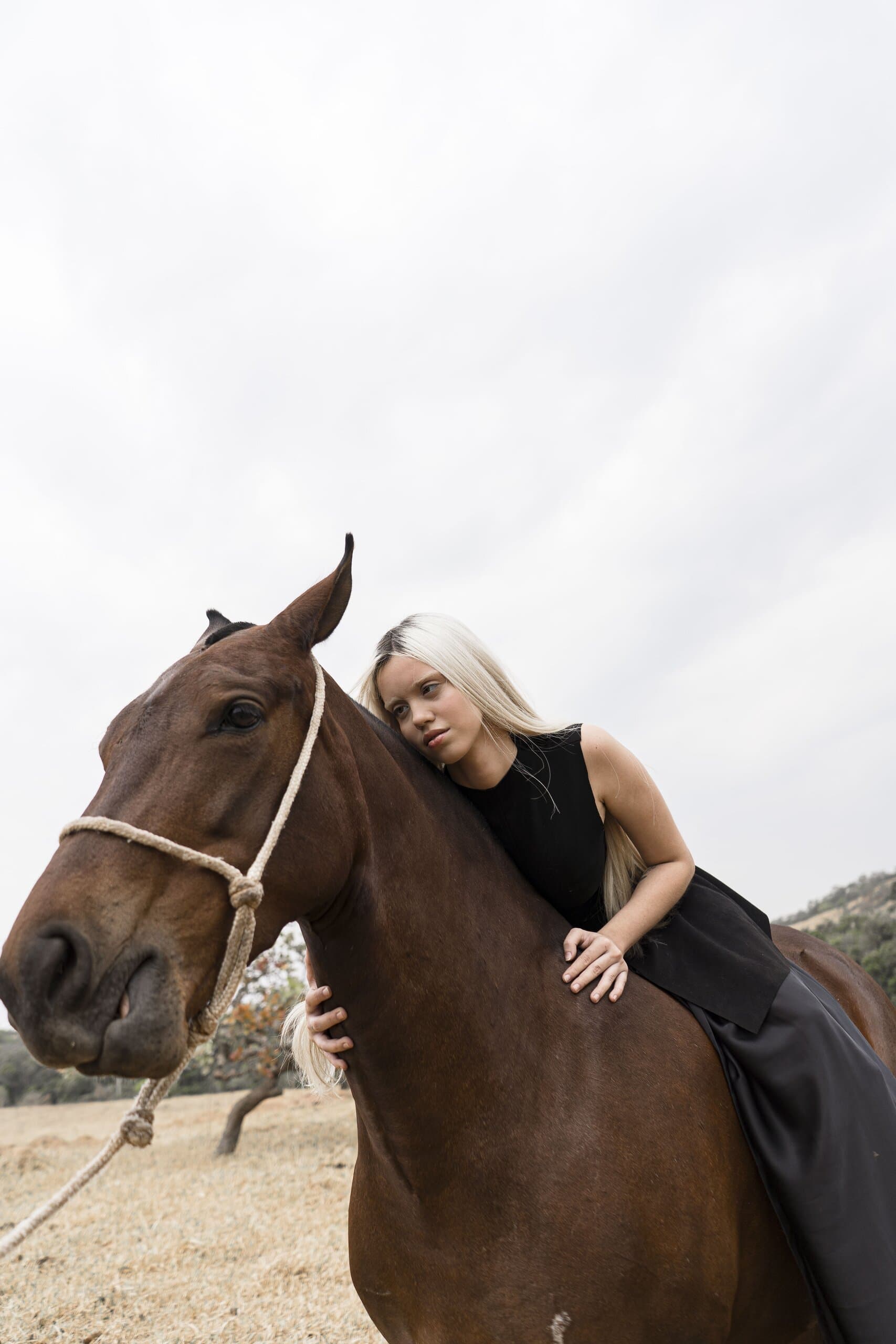 A blonde woman in black leans along the neck of a chestnut horse in a rope halter.