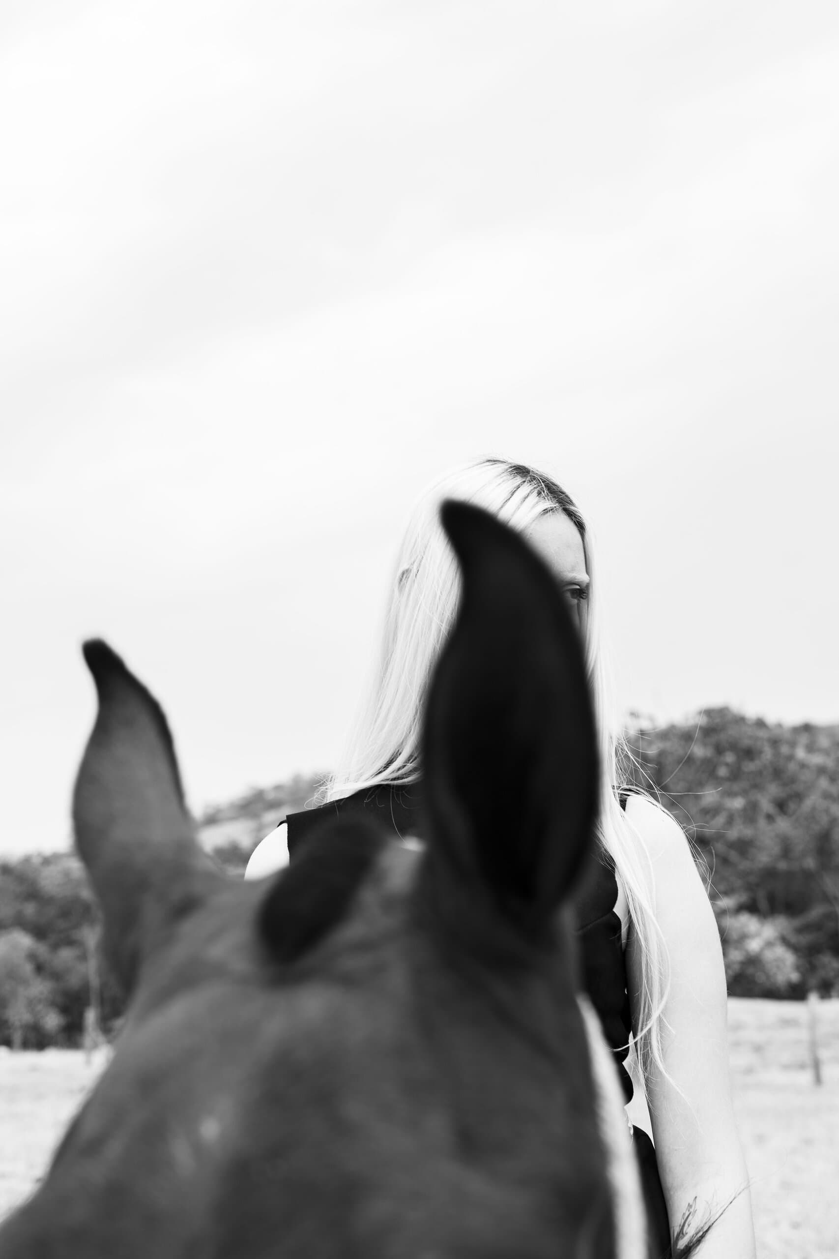 Black and white: a horse's ears blur in the foreground, a platinum-haired woman in black behind them.