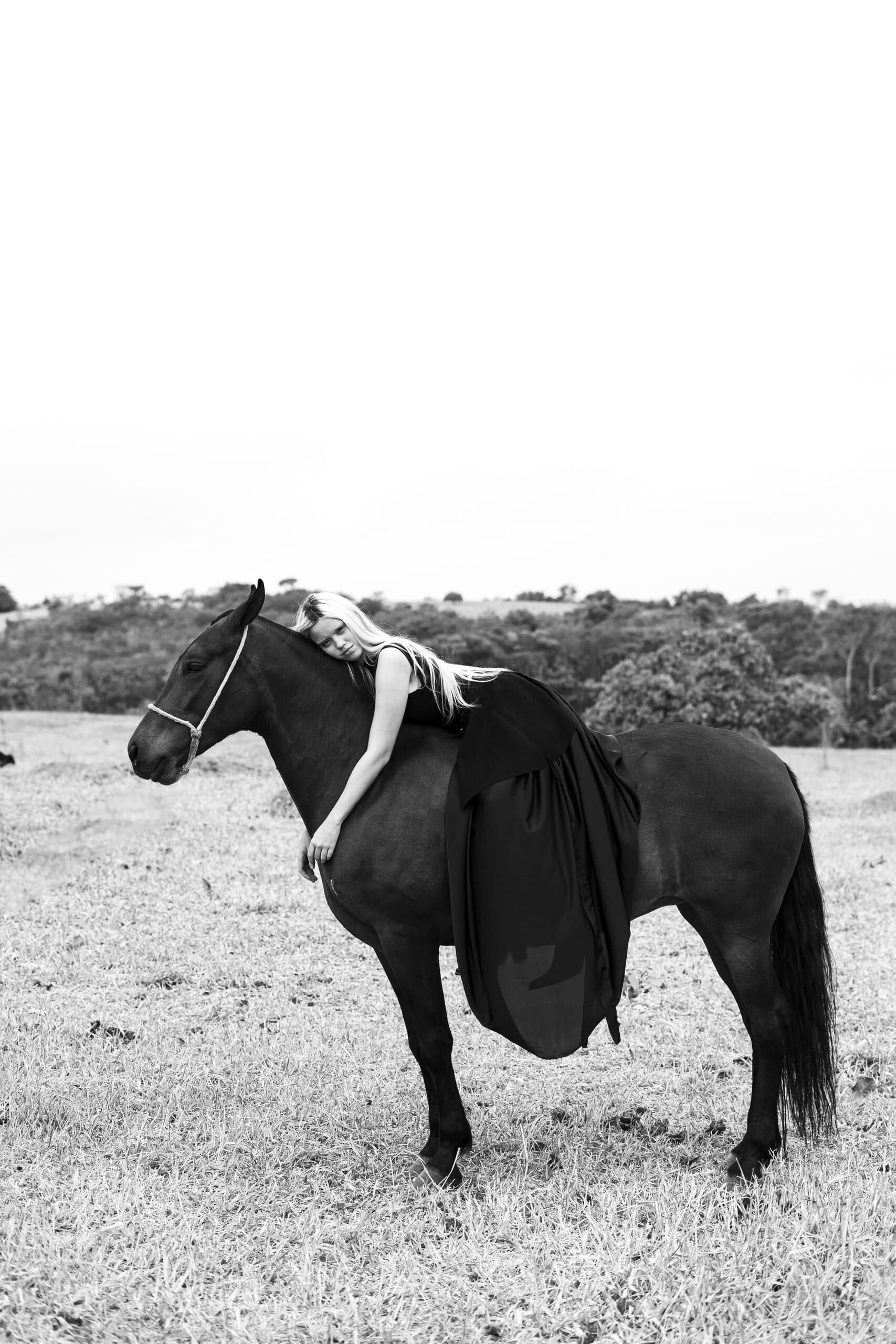 Black and white: a woman in a long black dress lies across the back of a dark horse in an open field.