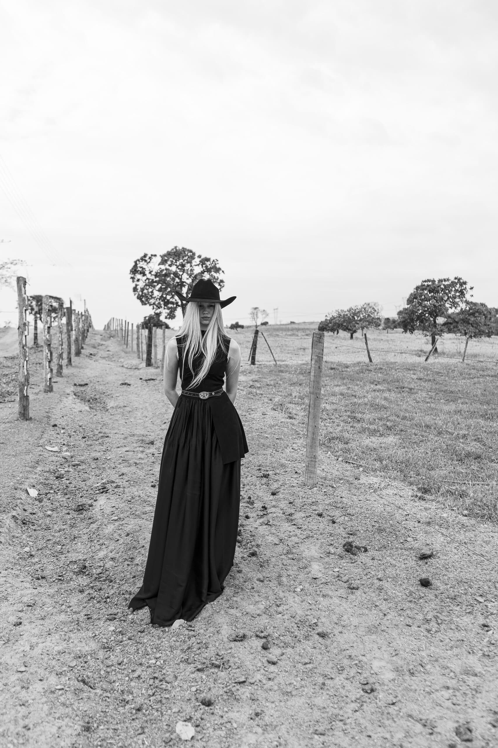 Black and white: a woman in a long black dress and cowboy hat stands between fence posts on a dirt track.