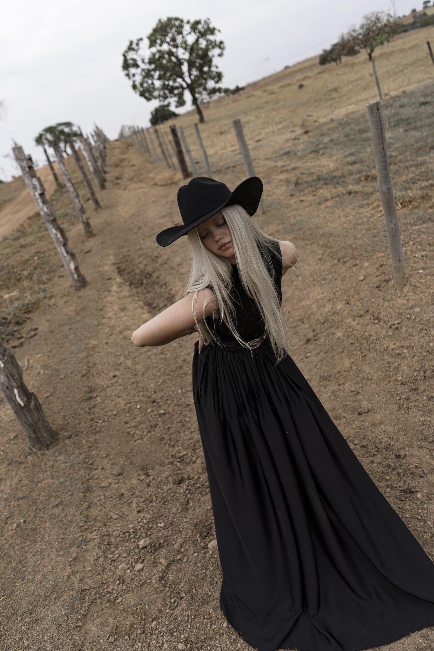 A woman in a black cowboy hat and flowing black gown turns on a sloping dirt track lined with fence posts.