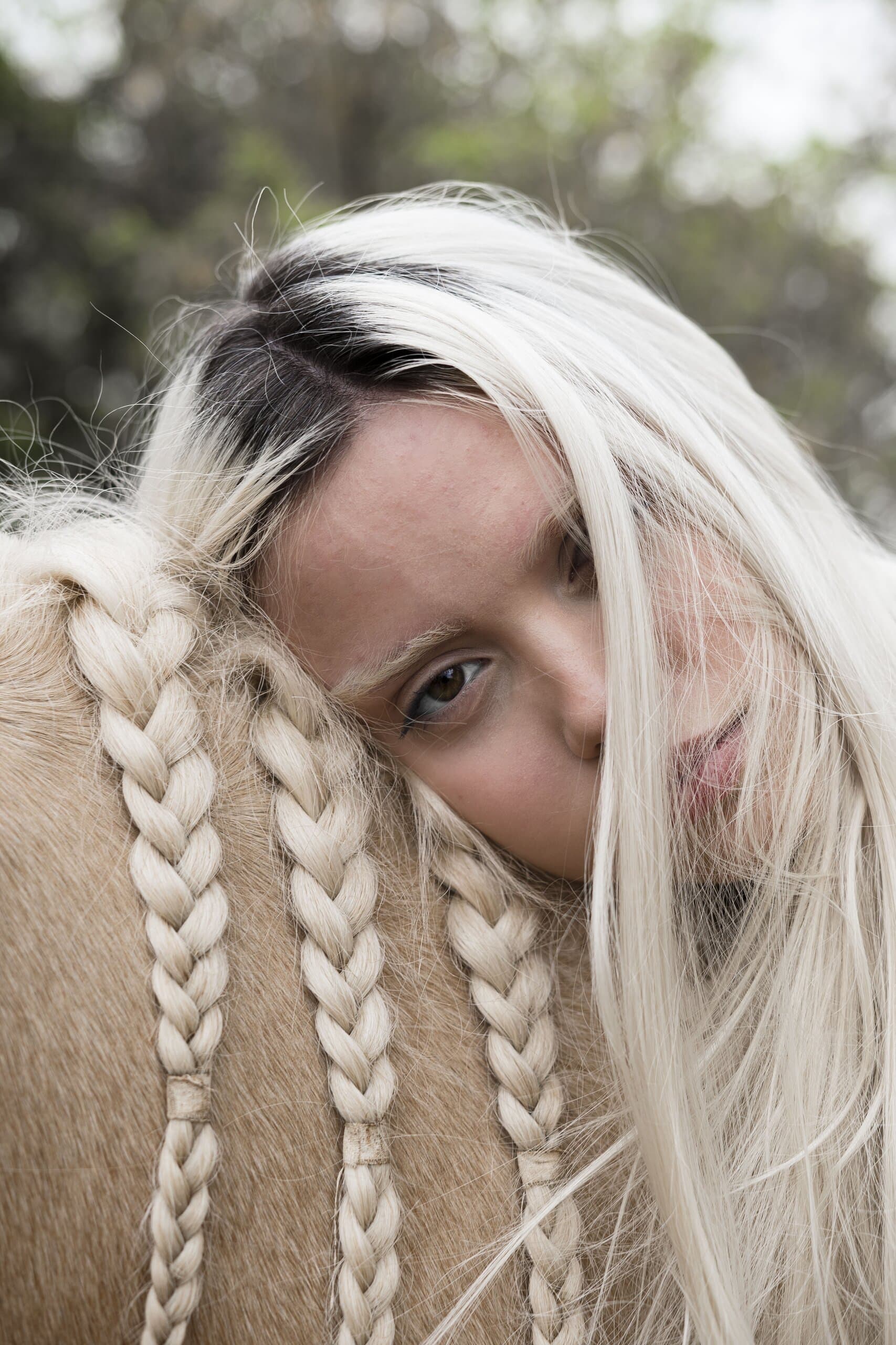 Close up: a woman with platinum hair rests her cheek on a palomino horse's braided mane.