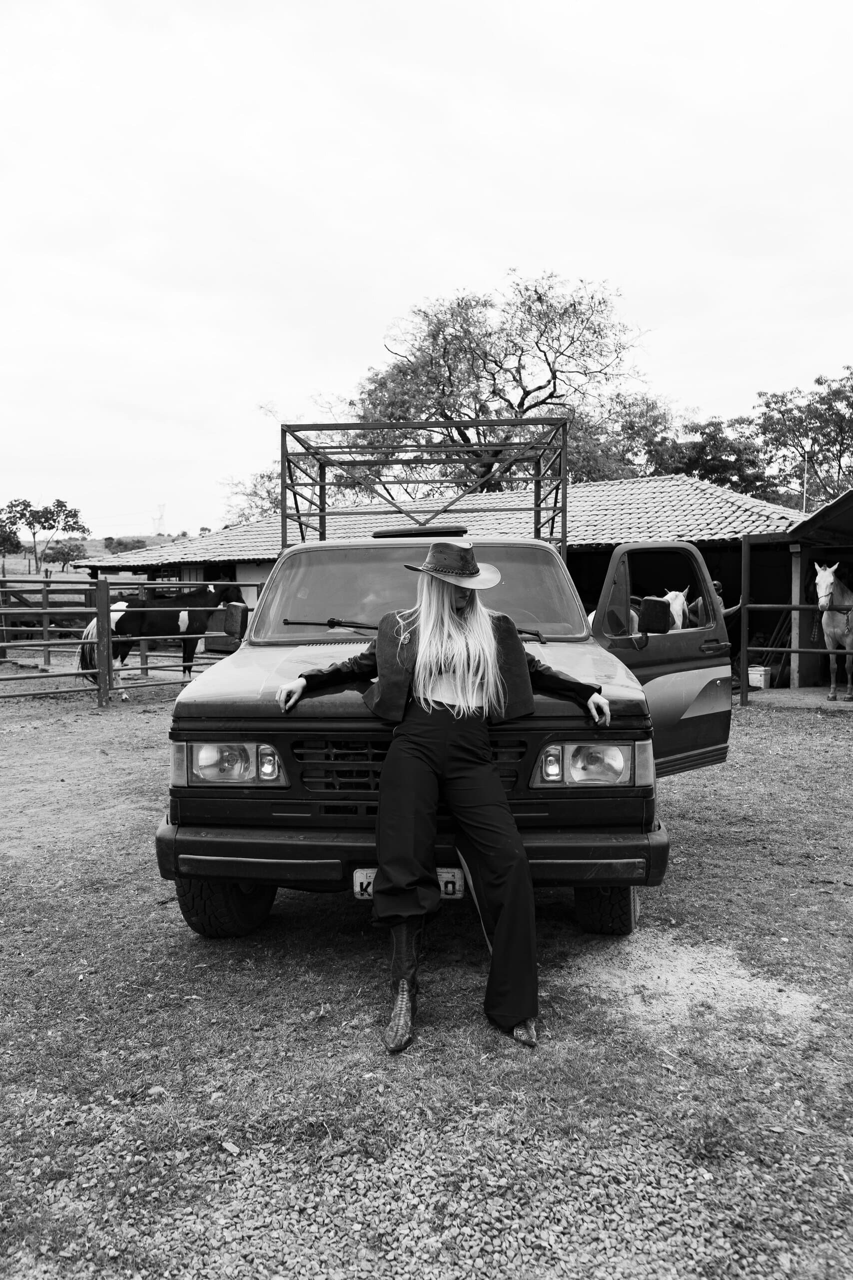 Black and white: a woman in a cowboy hat leans back on the bonnet of an old pick-up truck by a corral.