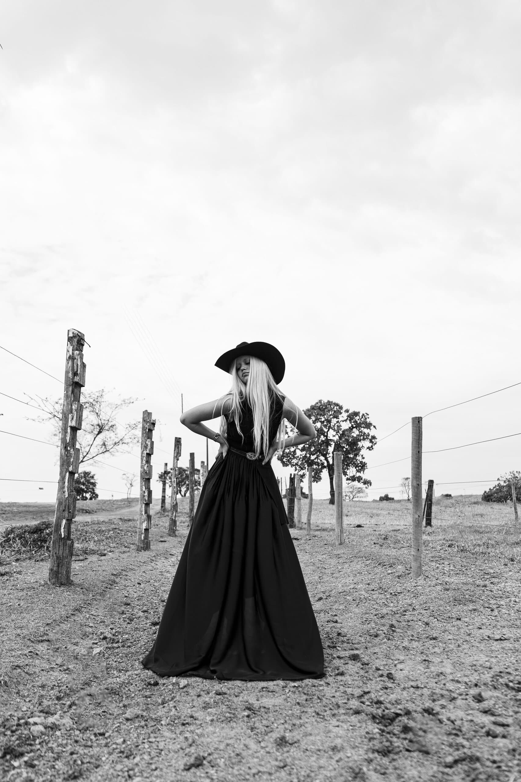 Black and white: hands on hips, a woman in a black hat and long dress stands among fence posts and wire.