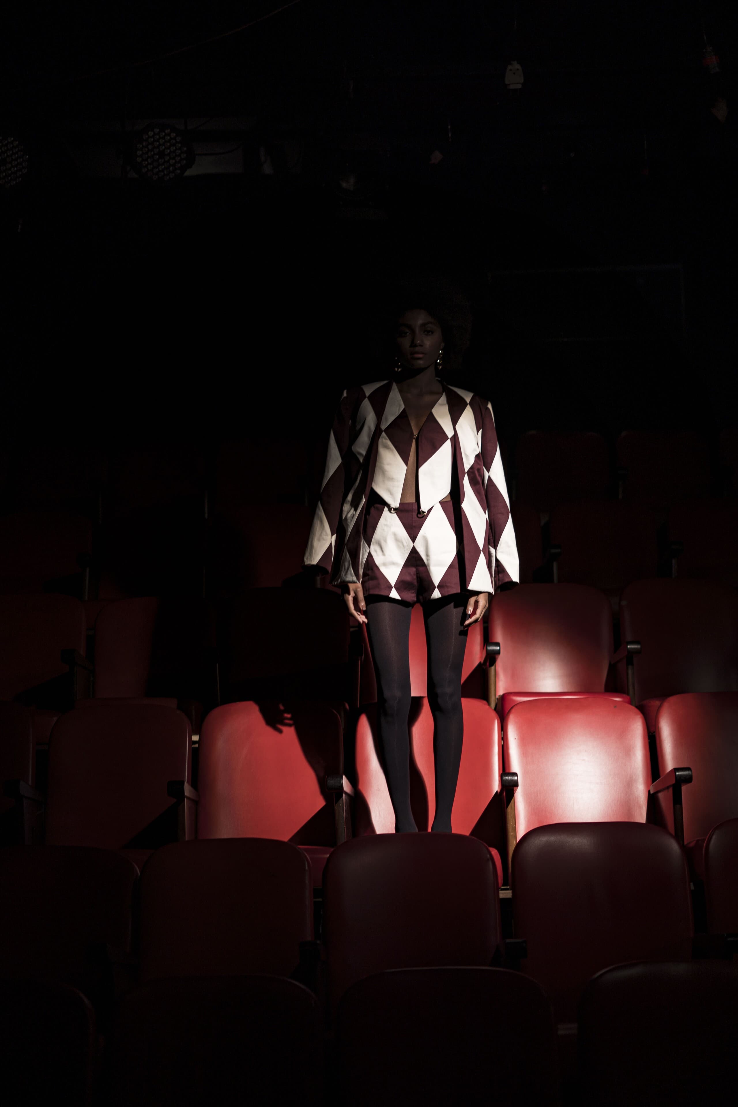 A figure in a harlequin top stands among rows of red seats in the dark.