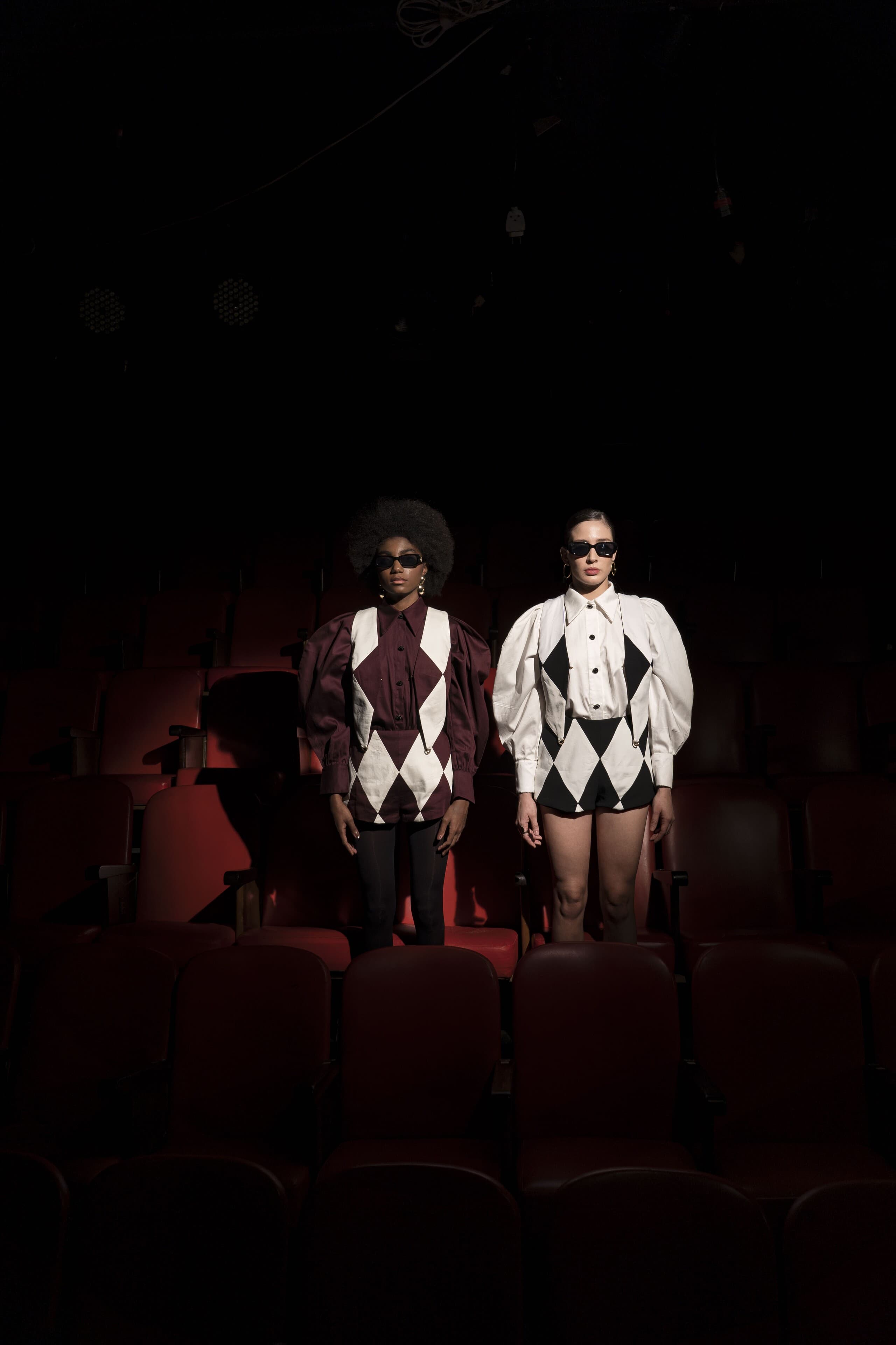 Two figures in harlequin tops stand side by side among the red seats.