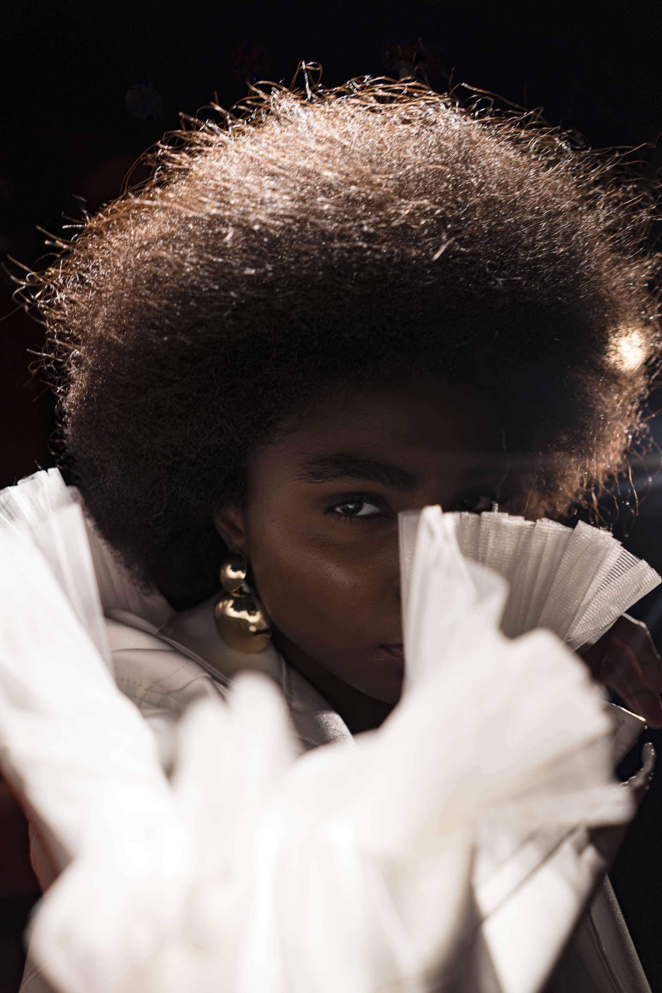 Close up: a face half hidden behind a ruffled white sleeve, a gold earring catching the light.