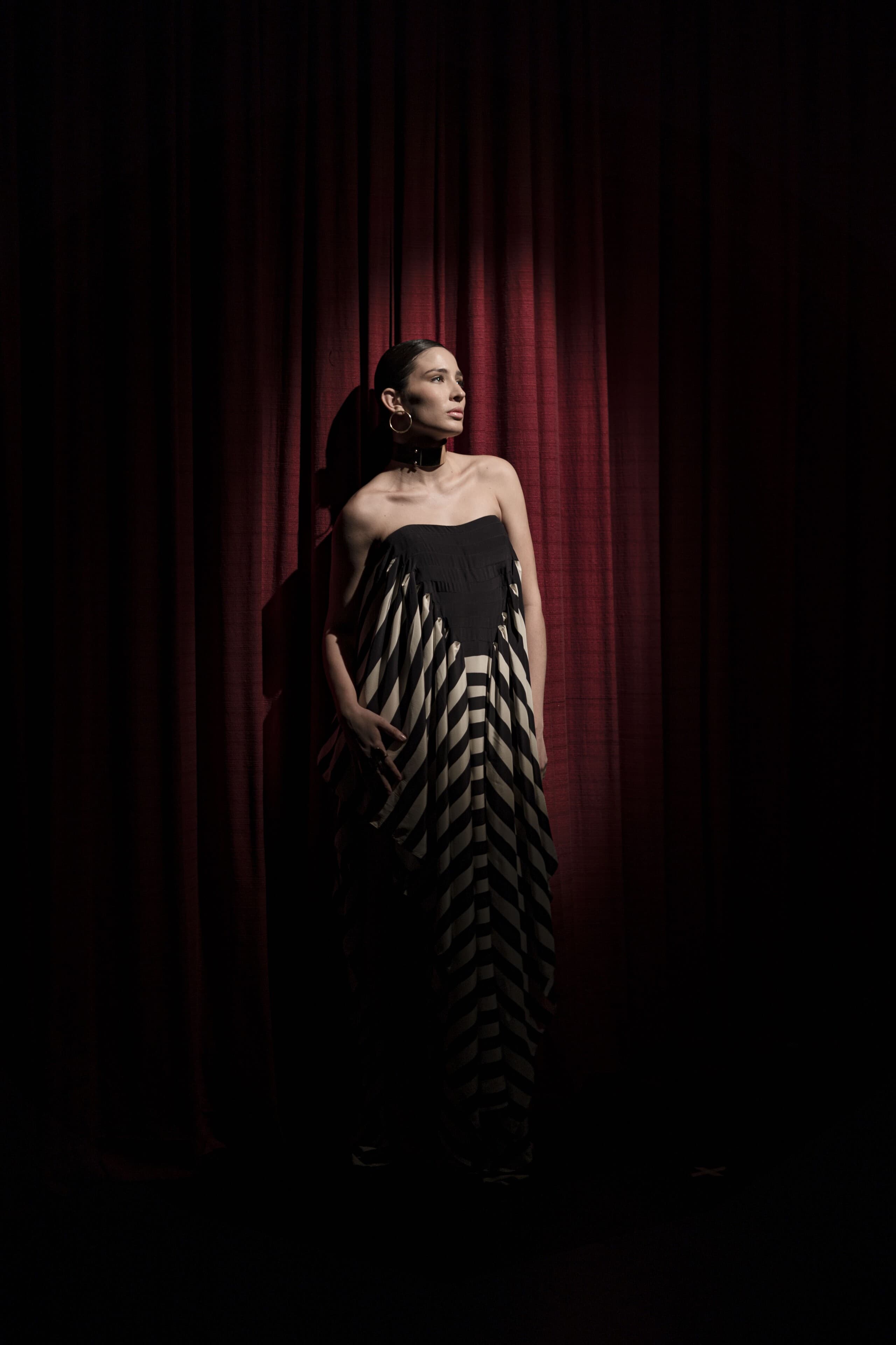 A woman in a striped gown stands before a deep red stage curtain.