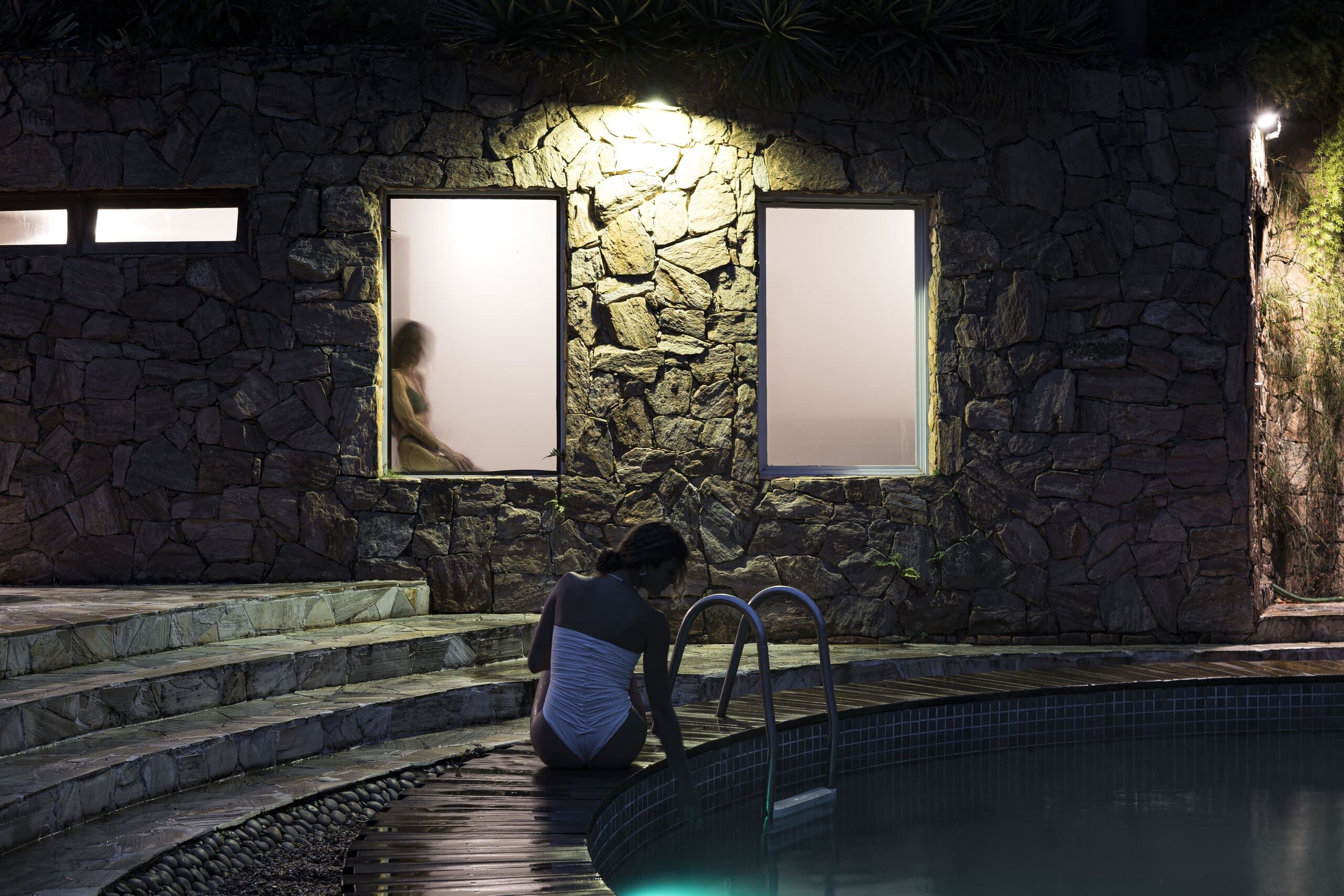 À noite, uma mulher sentada na borda de uma piscina sob um muro de pedra iluminado com janelas foscas.