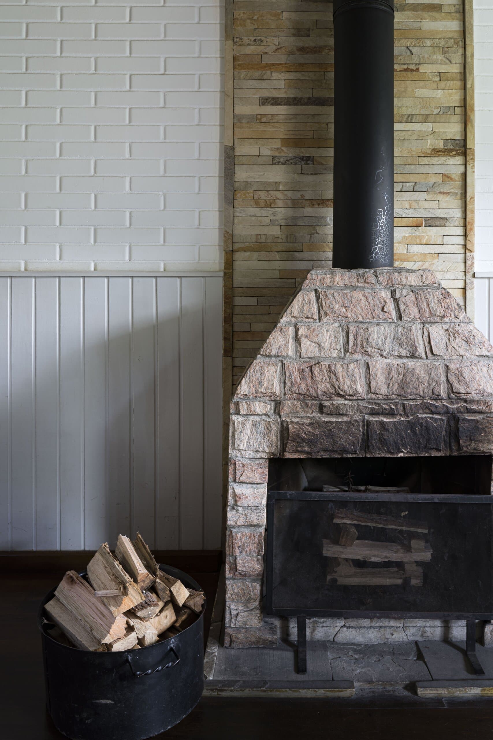 Uma lareira de pedra com chaminé preta contra tijolos brancos, um cesto de lenha ao lado.