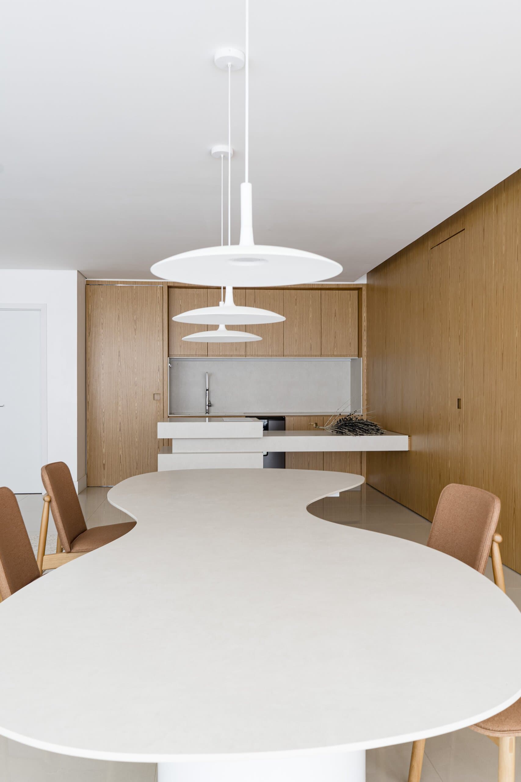 A curving white table runs towards the kitchen beneath stacked white pendants, timber chairs at its edge and a timber wall alongside.