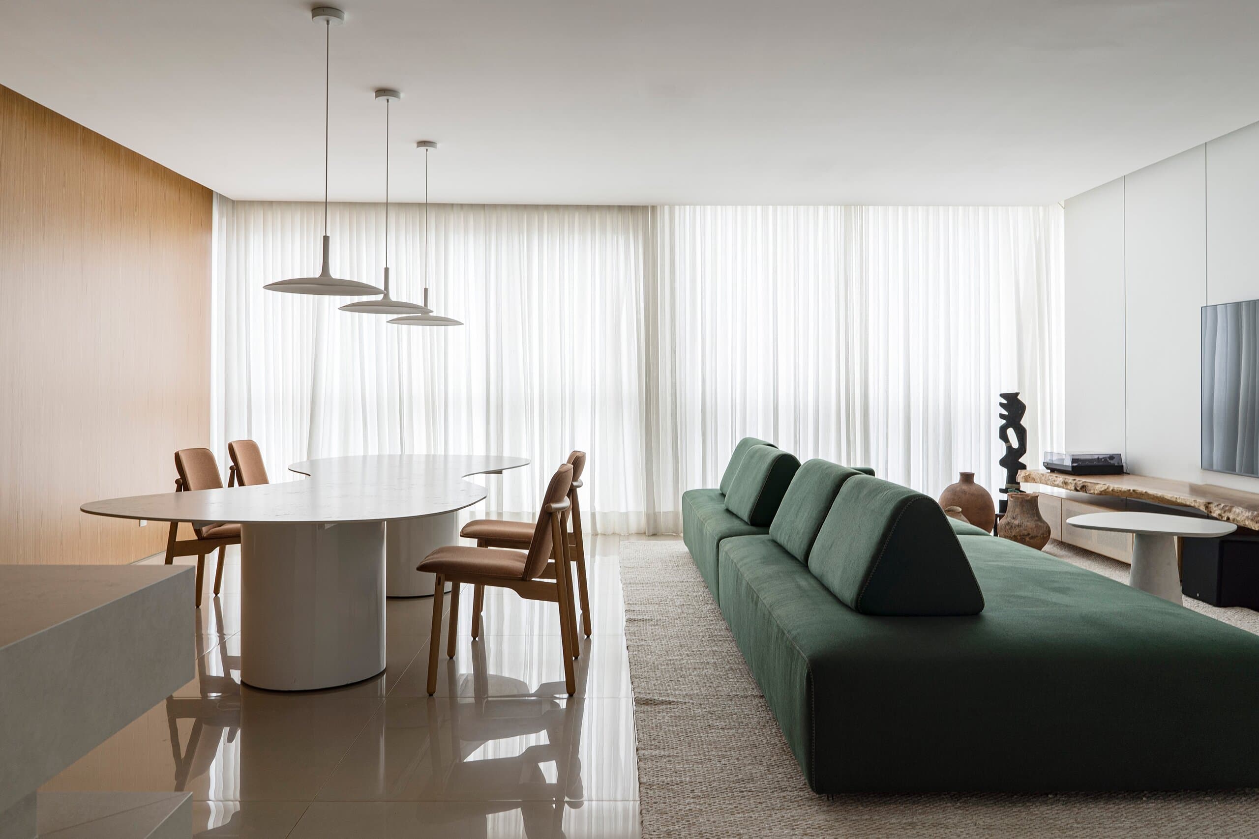 The dining table and long green sofa in soft light from sheer curtains, a dark carved sculpture on a timber console.