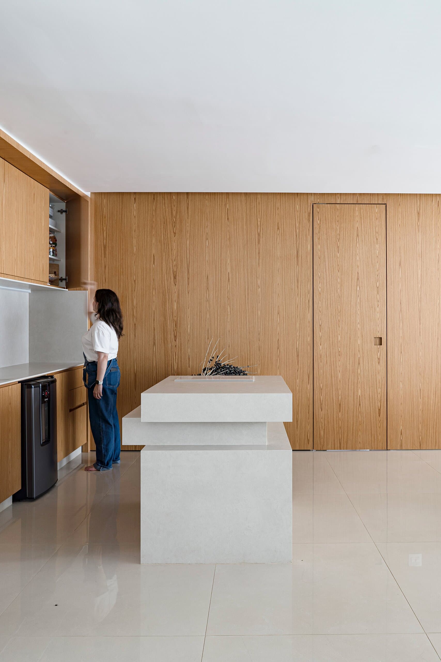 A woman reaches into a timber-clad kitchen wall beside a pale stone island, a flush timber door to one side.
