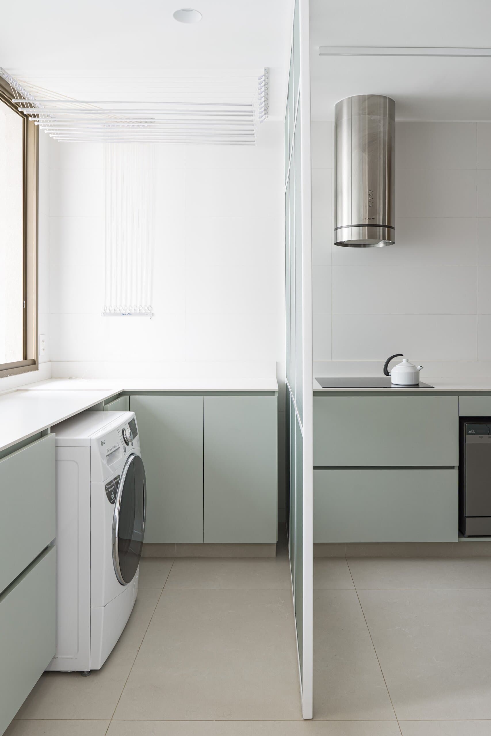 A sage-green kitchen behind a glass partition: a washing machine, a cylindrical steel hood and a kettle on the counter.