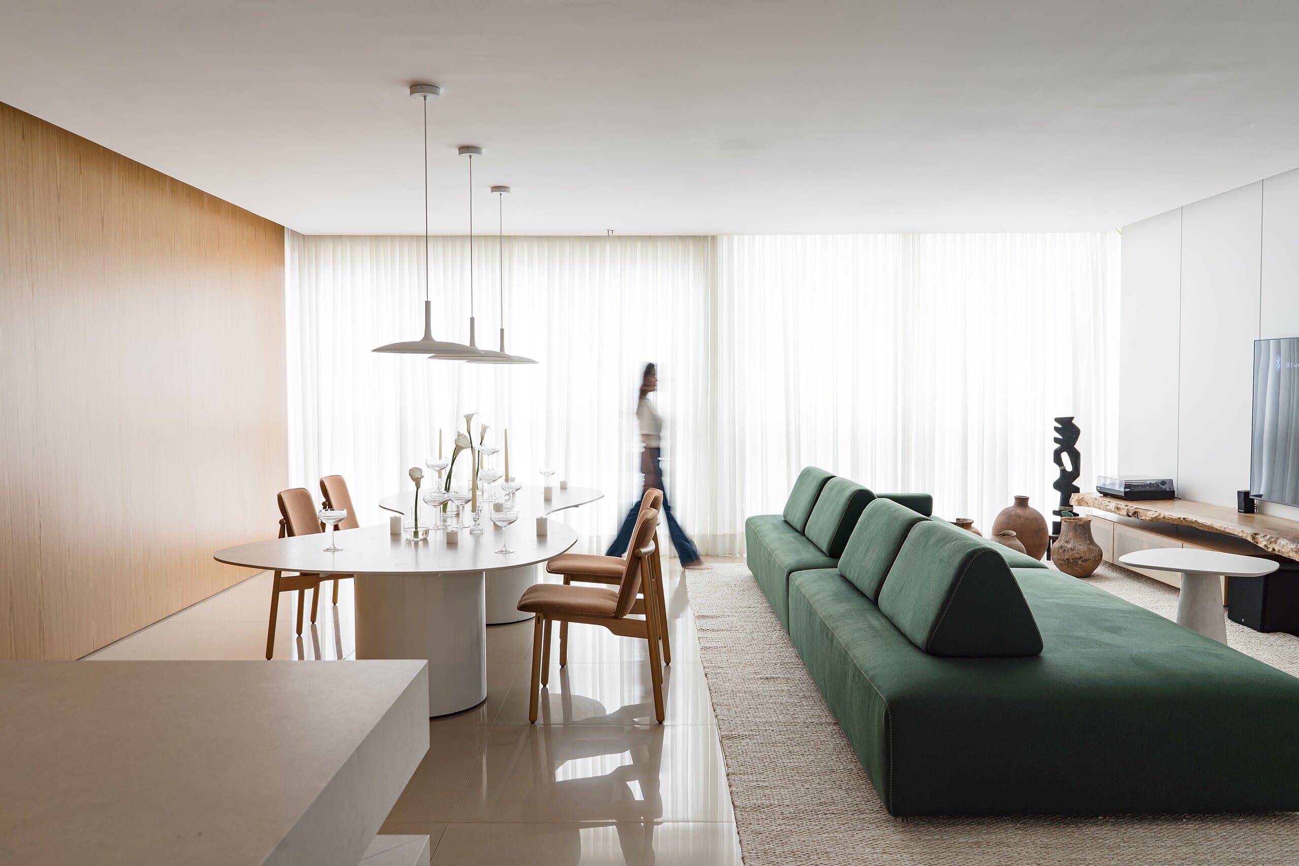 The open living and dining room: a woman walks past the oval table set with calla lilies, a long green sofa and timber-clad walls beneath three pendants.